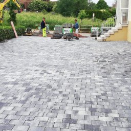 MARGELS SPÓŁKA Z OGRANICZONĄ ODPOWIEDZIALNOŚCIĄ - Ułożona kostka brukowa EHL na posesji, widoczni pracownicy i maszyny. W tle budynek z oknami i schodami. Na pierwszym planie piła do cięcia kostki.