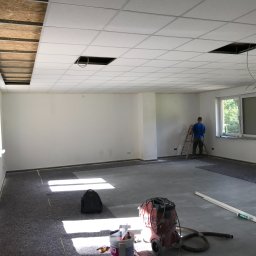 MARGELS SPÓŁKA Z OGRANICZONĄ ODPOWIEDZIALNOŚCIĄ - Wnętrze biura w trakcie remontu: białe ściany, podwieszany sufit z otworami, drabina i pracownik. Na podłodze folia, narzędzia i odkurzacz przemysłowy.