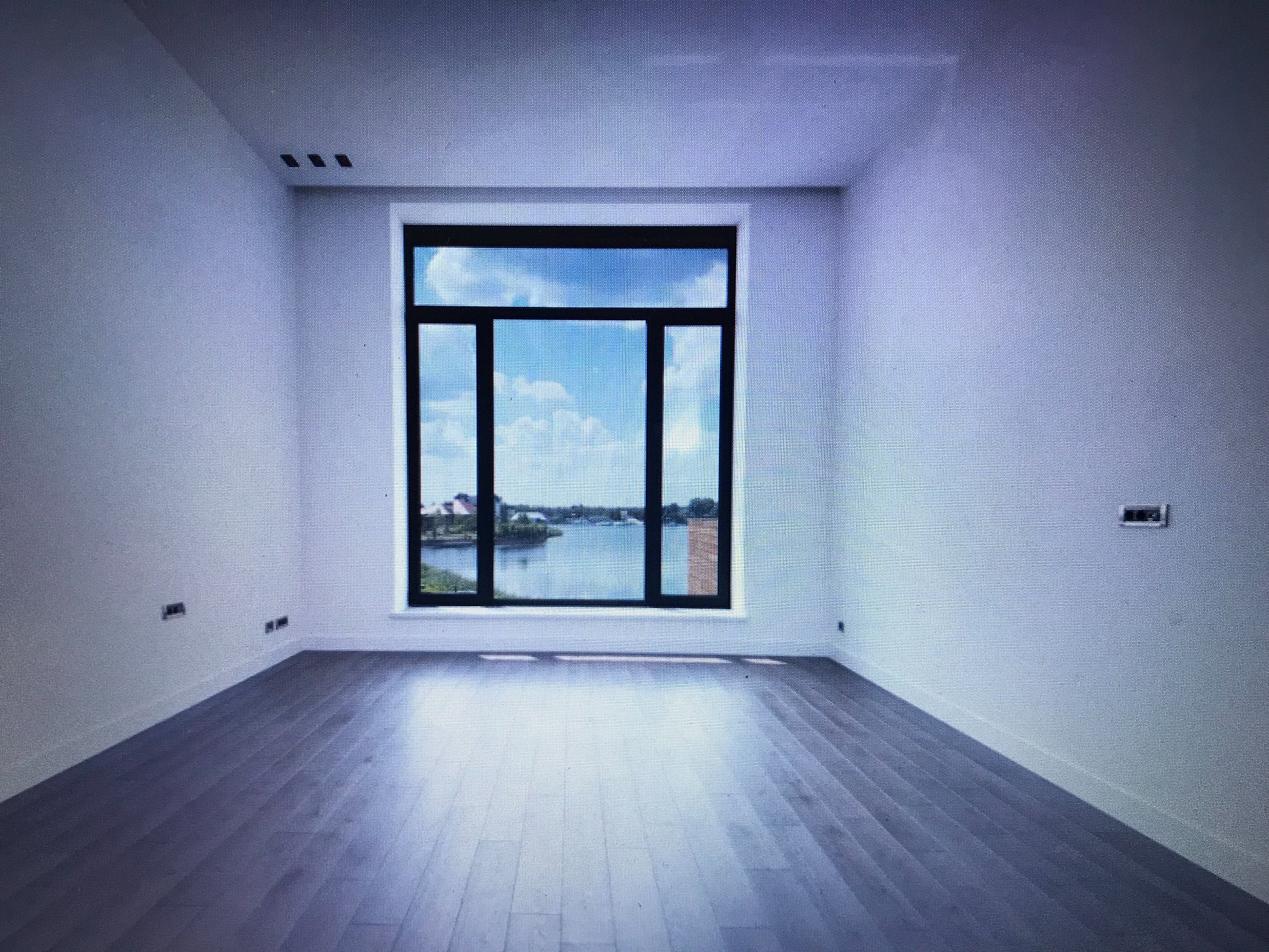 Puste, jasne wnętrze z drewnianą podłogą i dużym oknem z widokiem na wodę. Białe ściany z gniazdkami i wnękami na oświetlenie.