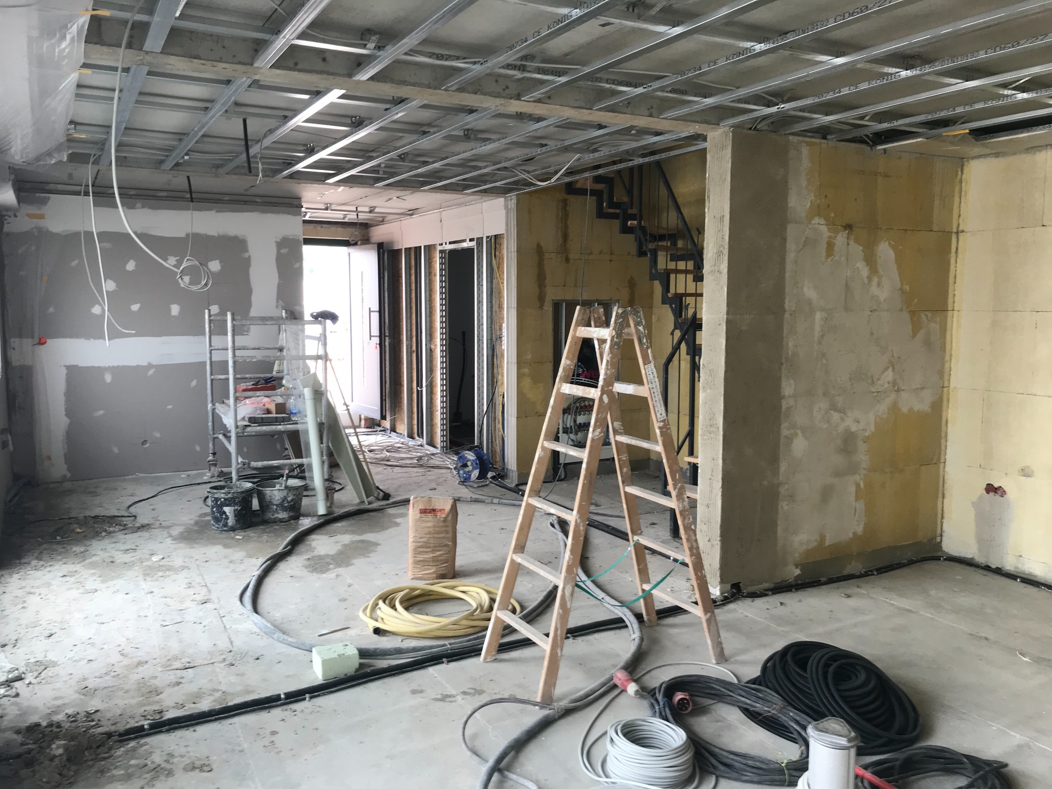 Wnętrze w trakcie remontu: widoczne rusztowanie sufitowe, ściany w trakcie szpachlowania, drabina, kable i worki z zaprawą. W tle schody i inne pomieszczenia.