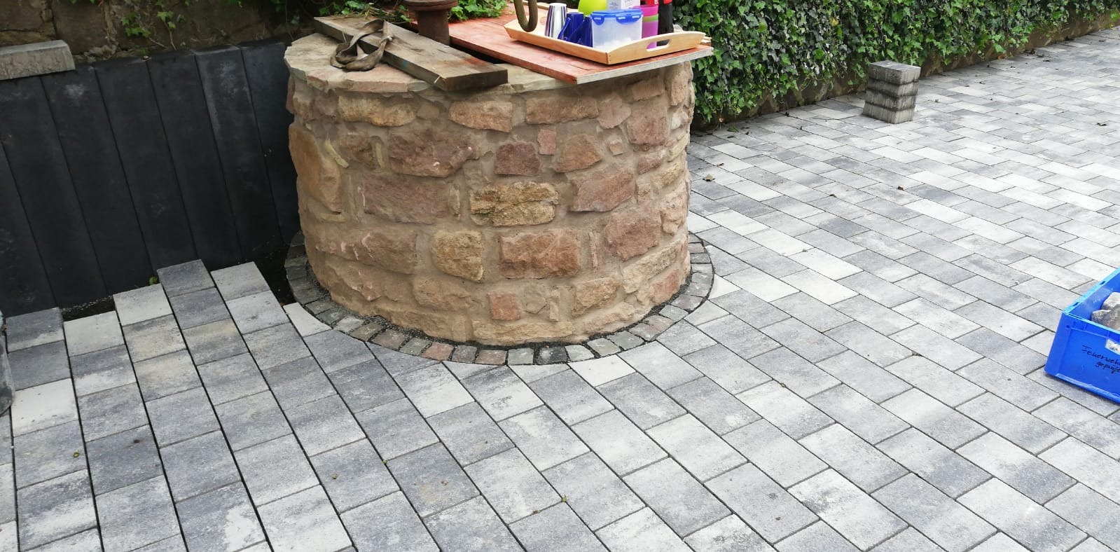 Studnia z kamienia otoczona szarą kostką brukową, schody z kostki prowadzą w dół. Zielona ściana z bluszczu w tle.