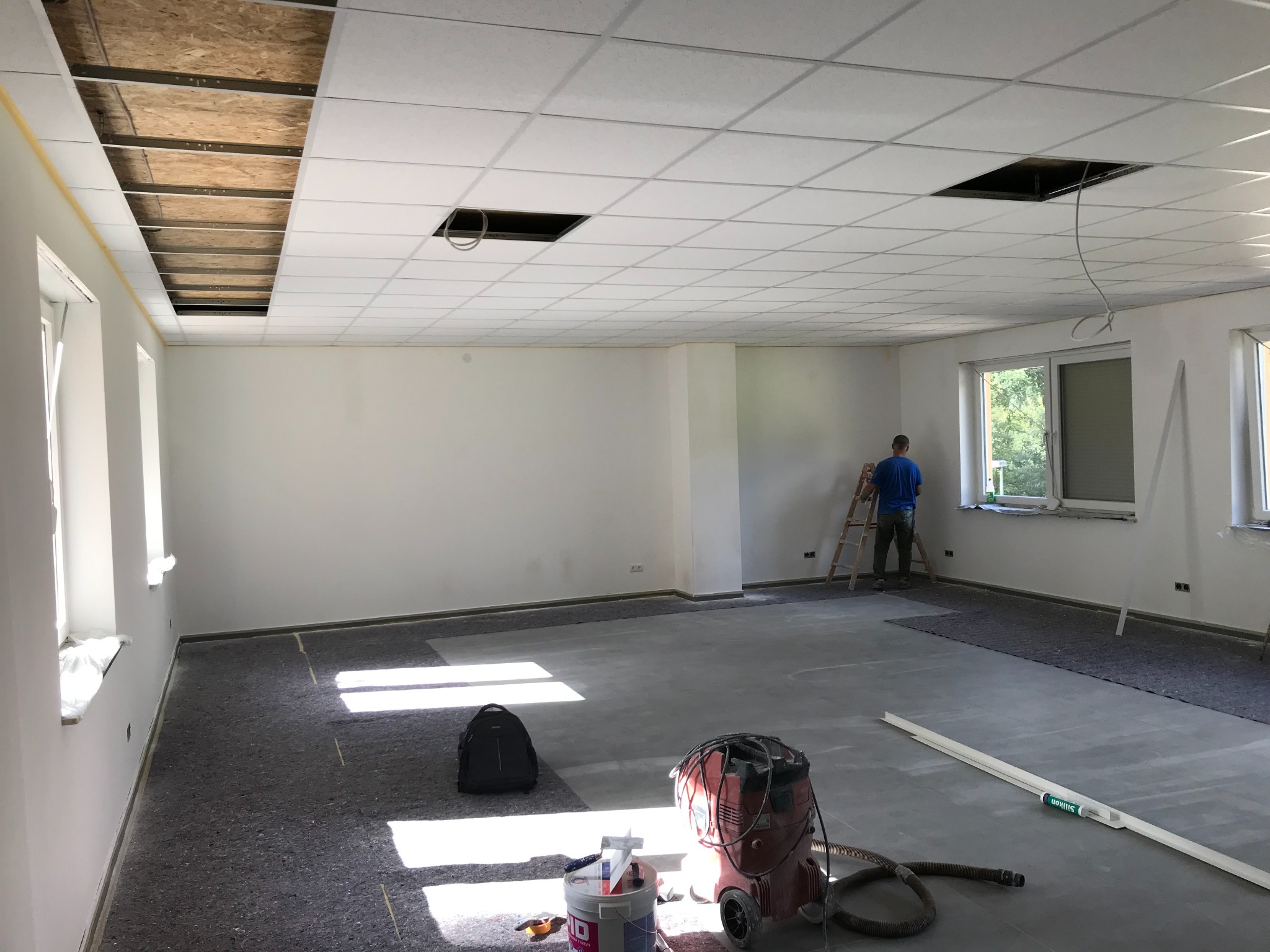 Wnętrze biura w trakcie remontu: białe ściany, podwieszany sufit z otworami, drabina i pracownik. Na podłodze folia, narzędzia i odkurzacz przemysłowy.