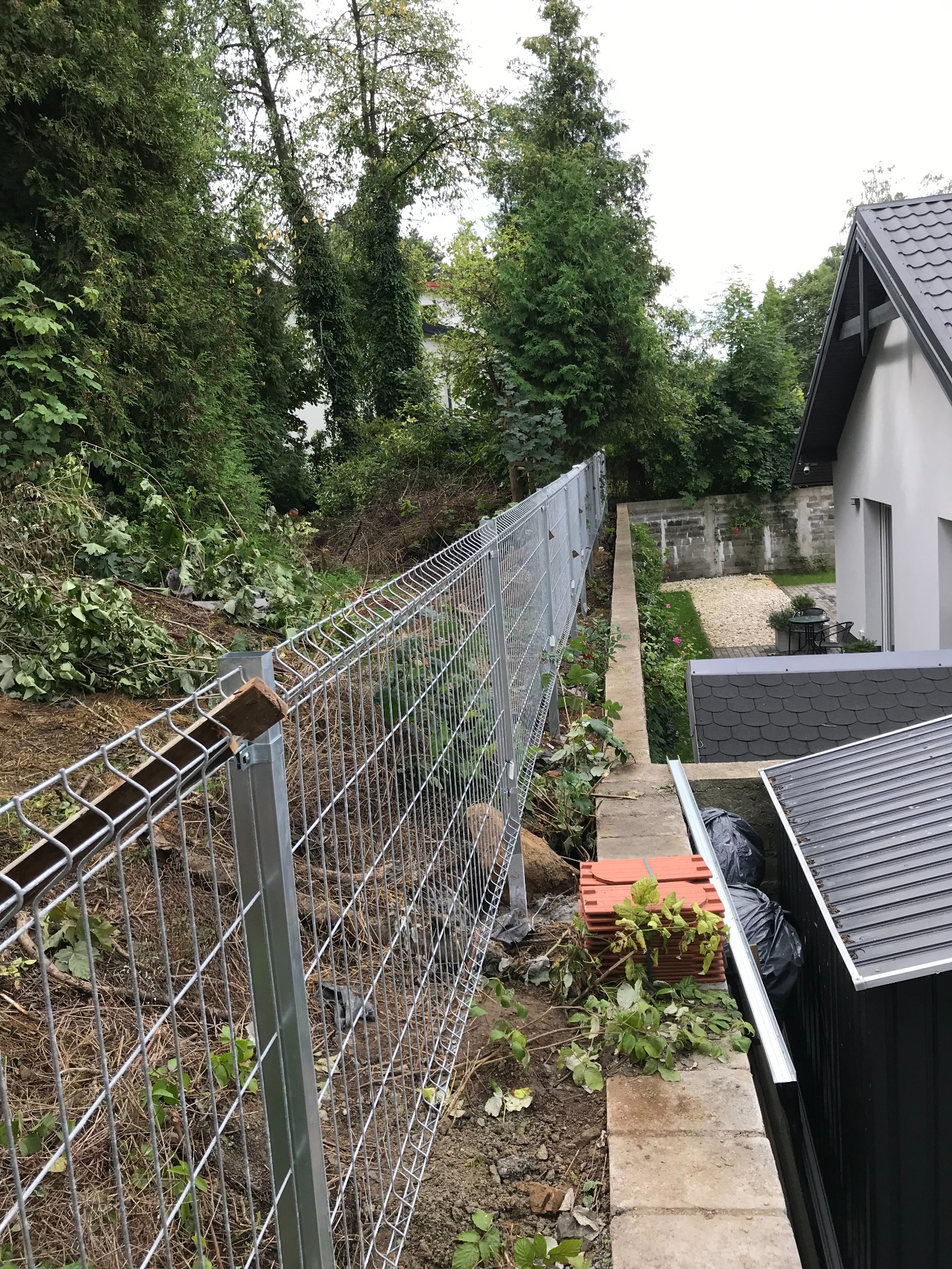 Montaż ogrodzenia panelowego na nierównym terenie, widok z boku, zieleń w tle. Elementy konstrukcyjne ogrodzenia widoczne w zbliżeniu, świeżo zamontowane.