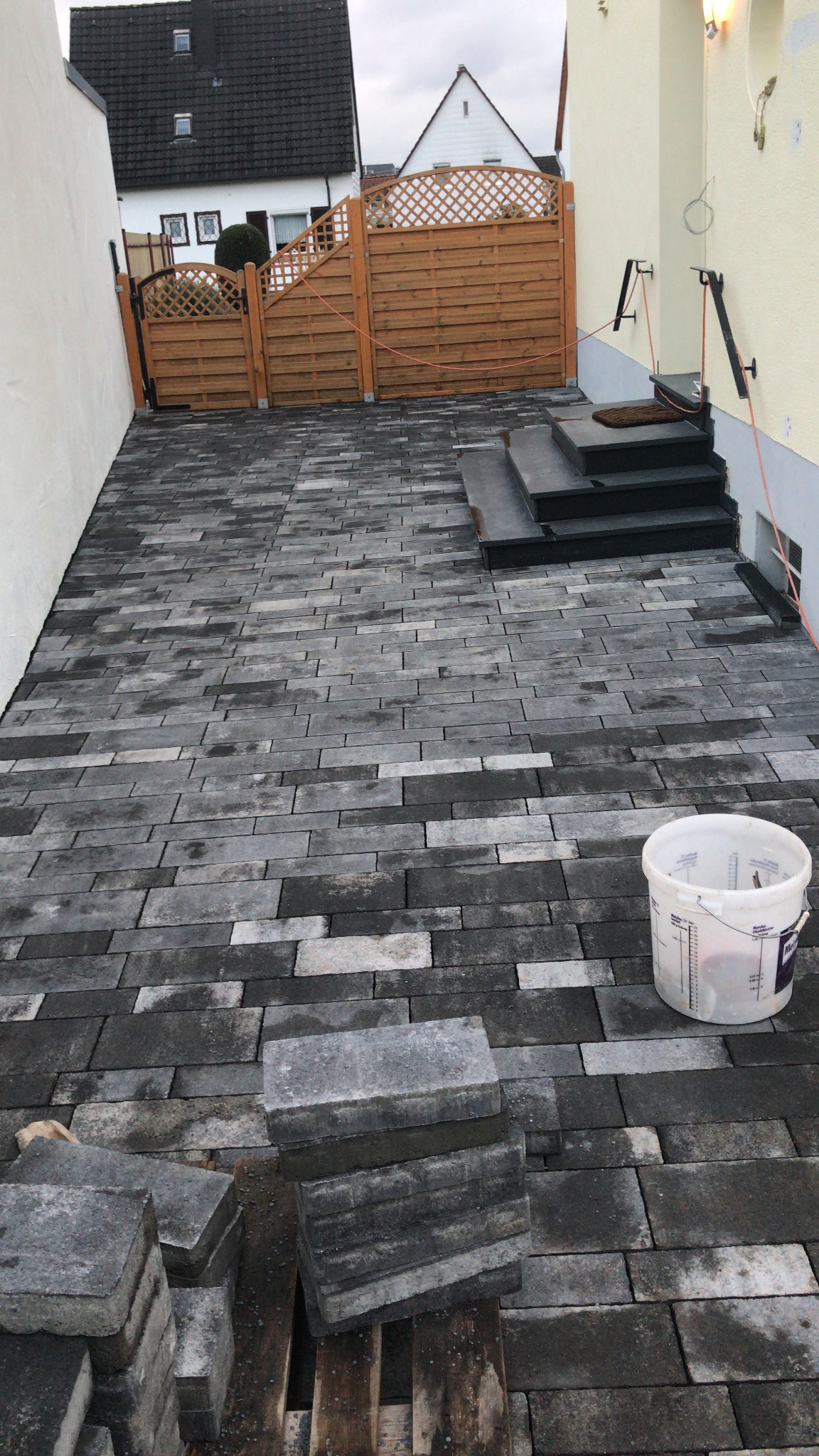 Układanie szarej kostki brukowej na podjeździe, widoczne niezakończone prace i wiadro. W tle drewniany płot i schody wejściowe do budynku.