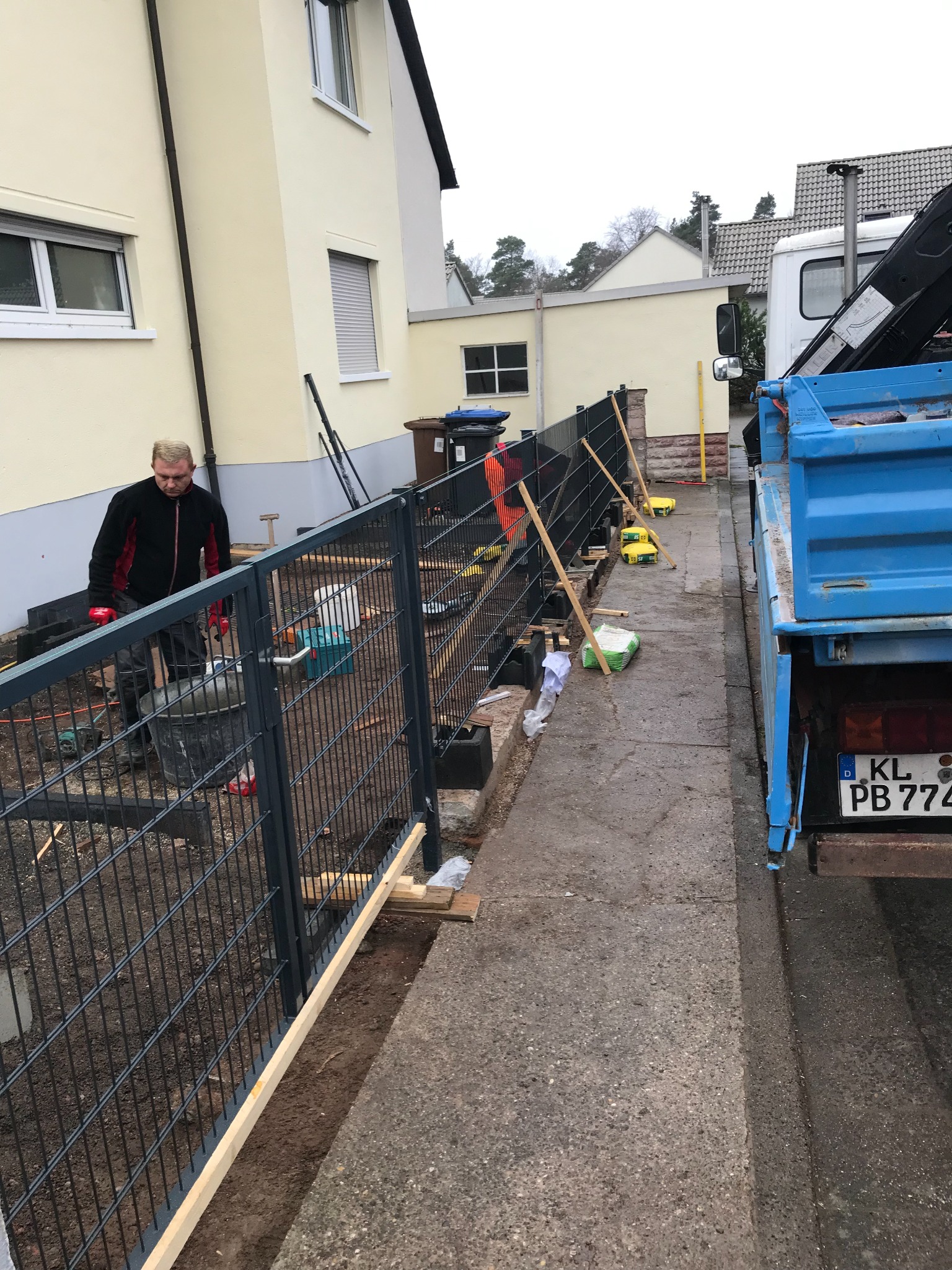 Montaż metalowego ogrodzenia panelowego przy domu jednorodzinnym. Widoczny pracownik, materiały budowlane i ciężarówka. Ujęcie z boku, w trakcie prac.