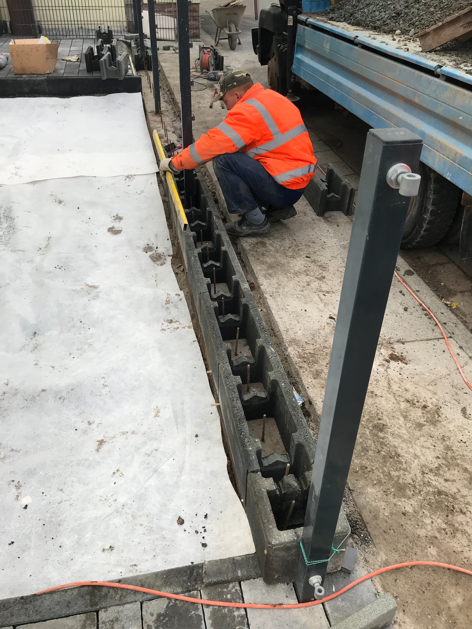 Pracownik w pomarańczowej kurtce poziomuje czarną obrzeże zbrojoną do wylewki fundamentu pod ogrodzenie, na ziemi rozłożona biała geowłóknina.