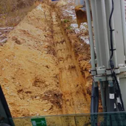 Wąski wykop w ziemi, widok z kabiny koparki, z boku elementy maszyny. W tle drzewa i niebieskie rury. Ślady śniegu na ziemi.