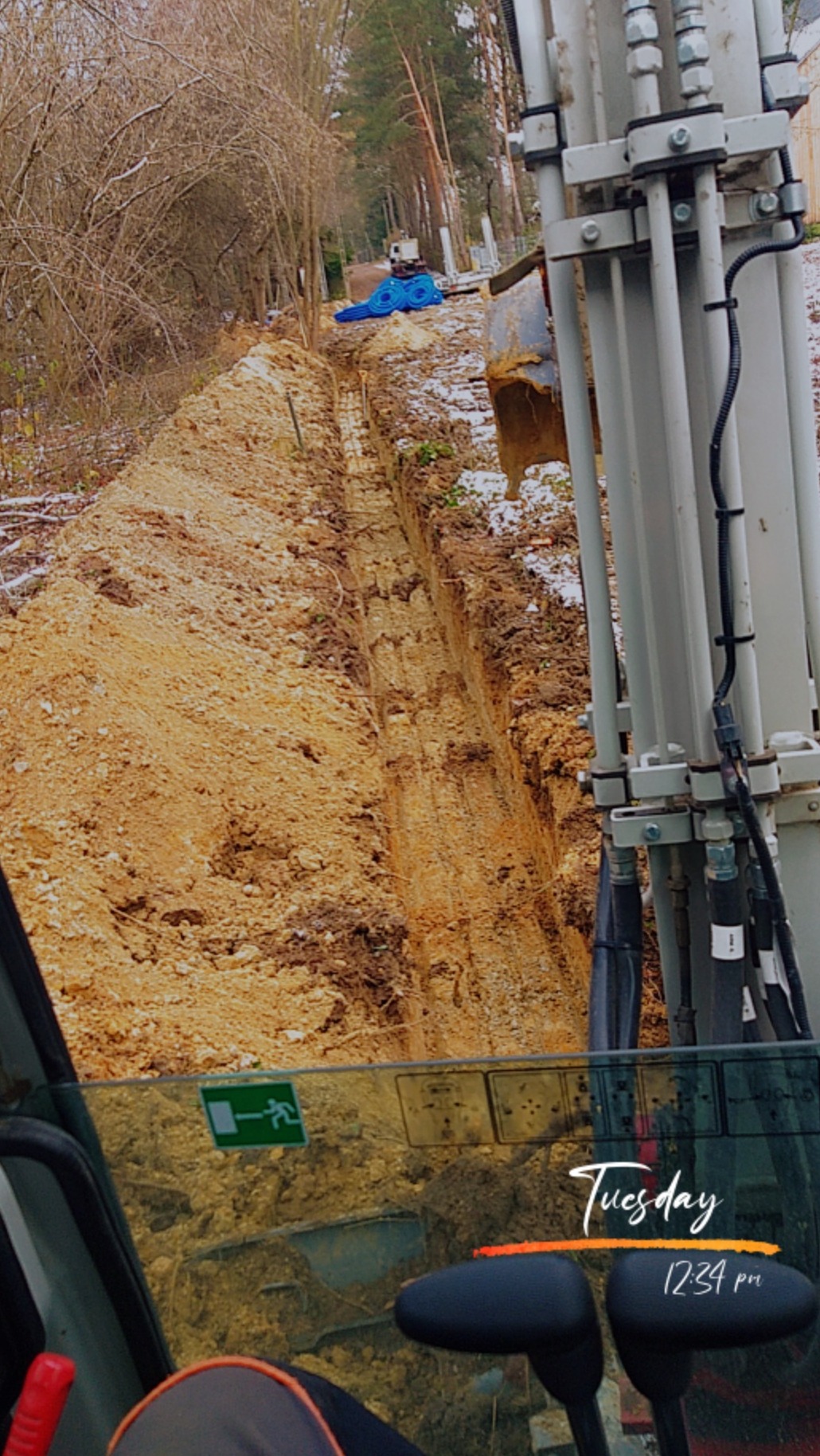 Wąski wykop w ziemi, widok z kabiny koparki, z boku elementy maszyny. W tle drzewa i niebieskie rury. Ślady śniegu na ziemi.