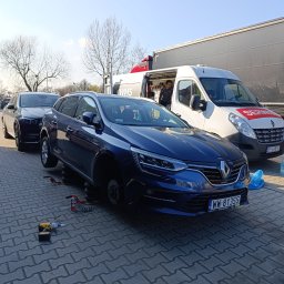 MOBILNA WULKANIZACJA WMS - Niebieskie kombi Renault bez koła, podnośnik i narzędzia obok. W tle bus serwisowy i ciężarówka na brukowanym placu. Usługa wymiany opon w terenie.