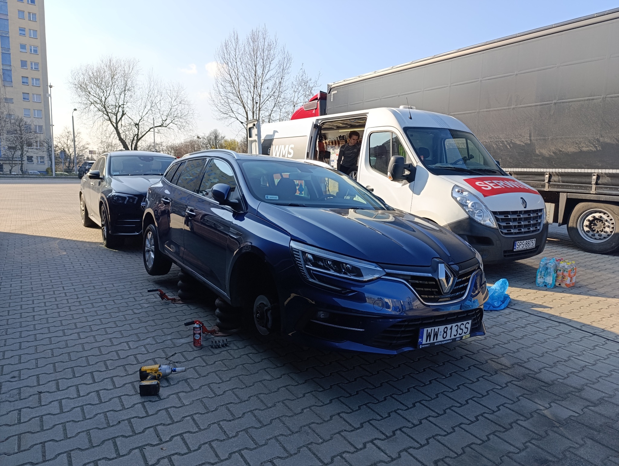 Niebieskie kombi Renault bez koła, podnośnik i narzędzia obok. W tle bus serwisowy i ciężarówka na brukowanym placu. Usługa wymiany opon w terenie.
