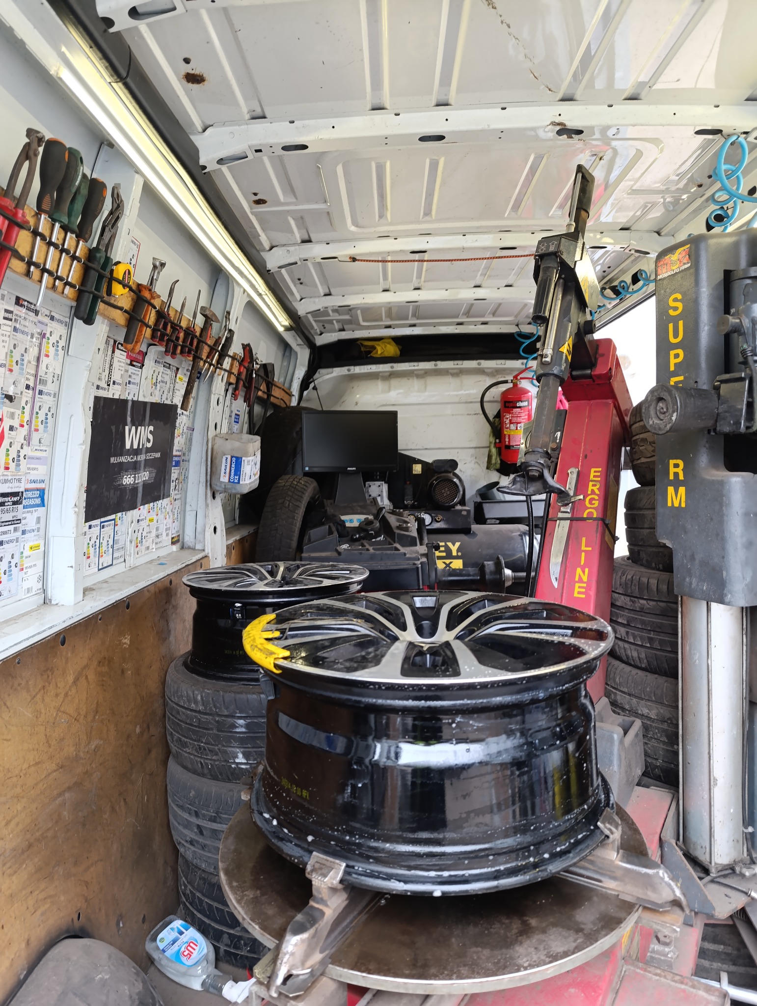 Wnętrze mobilnego warsztatu wulkanizacyjnego: narzędzia, opony, felgi na maszynie do wymiany opon. W tle widoczny monitor i gaśnica.