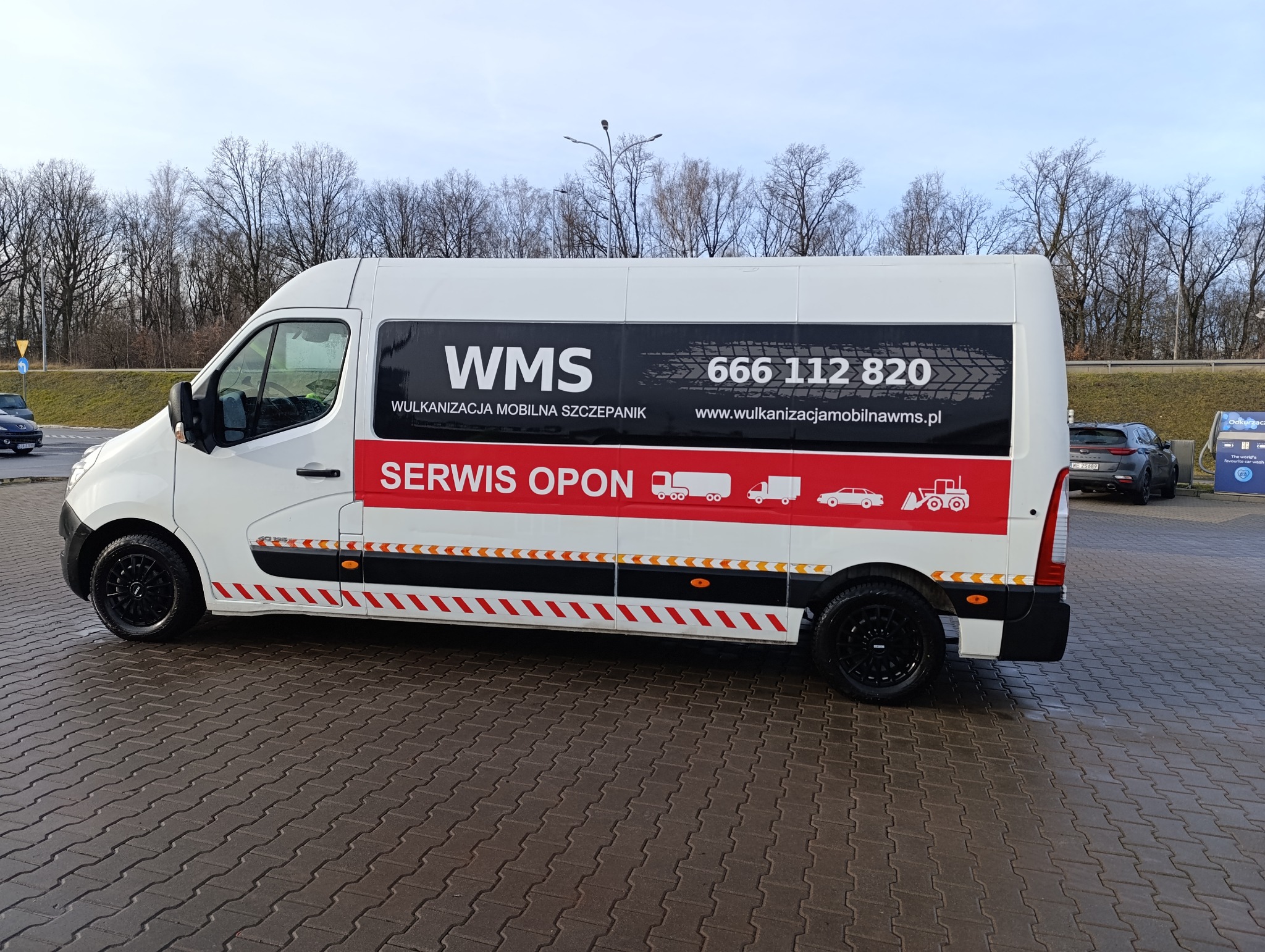 Mobilny serwis opon: biały bus z logo WMS, numerem telefonu i hasłem 'Wulkanizacja Mobilna Szczepanik'. Grafika z pojazdami na czerwonym pasie.