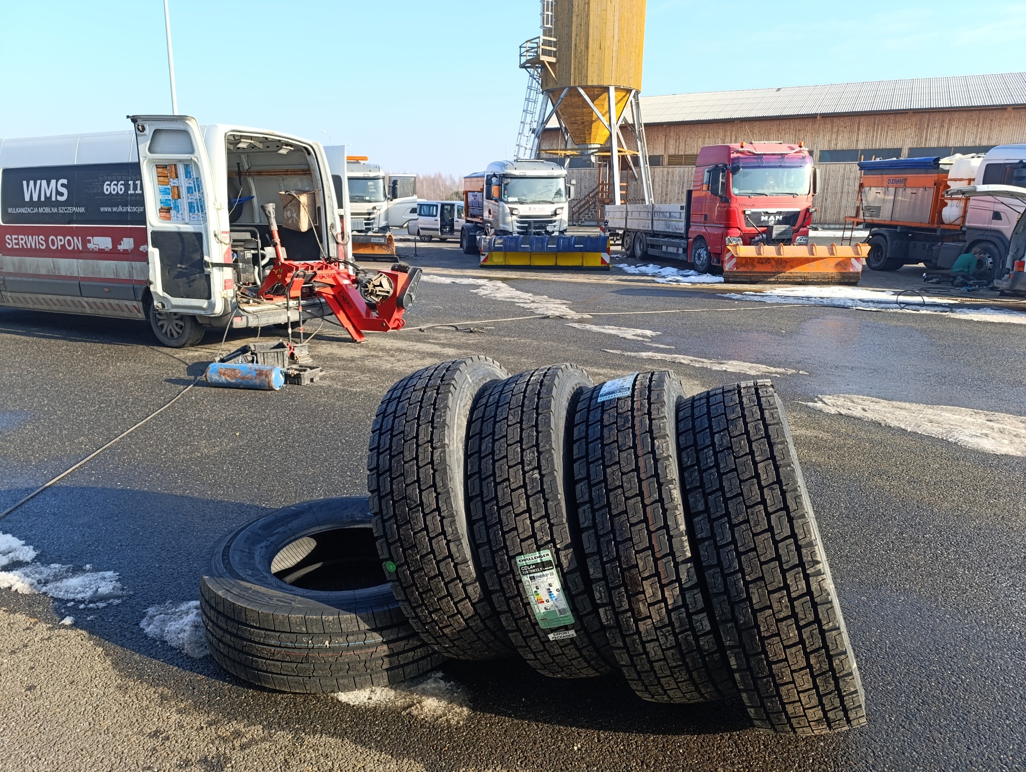 Mobilny serwis opon: opony ciężarowe z etykietami na tle wozu serwisowego WMS i sprzętu do wymiany opon. W tle zaparkowane ciężarówki z pługami.