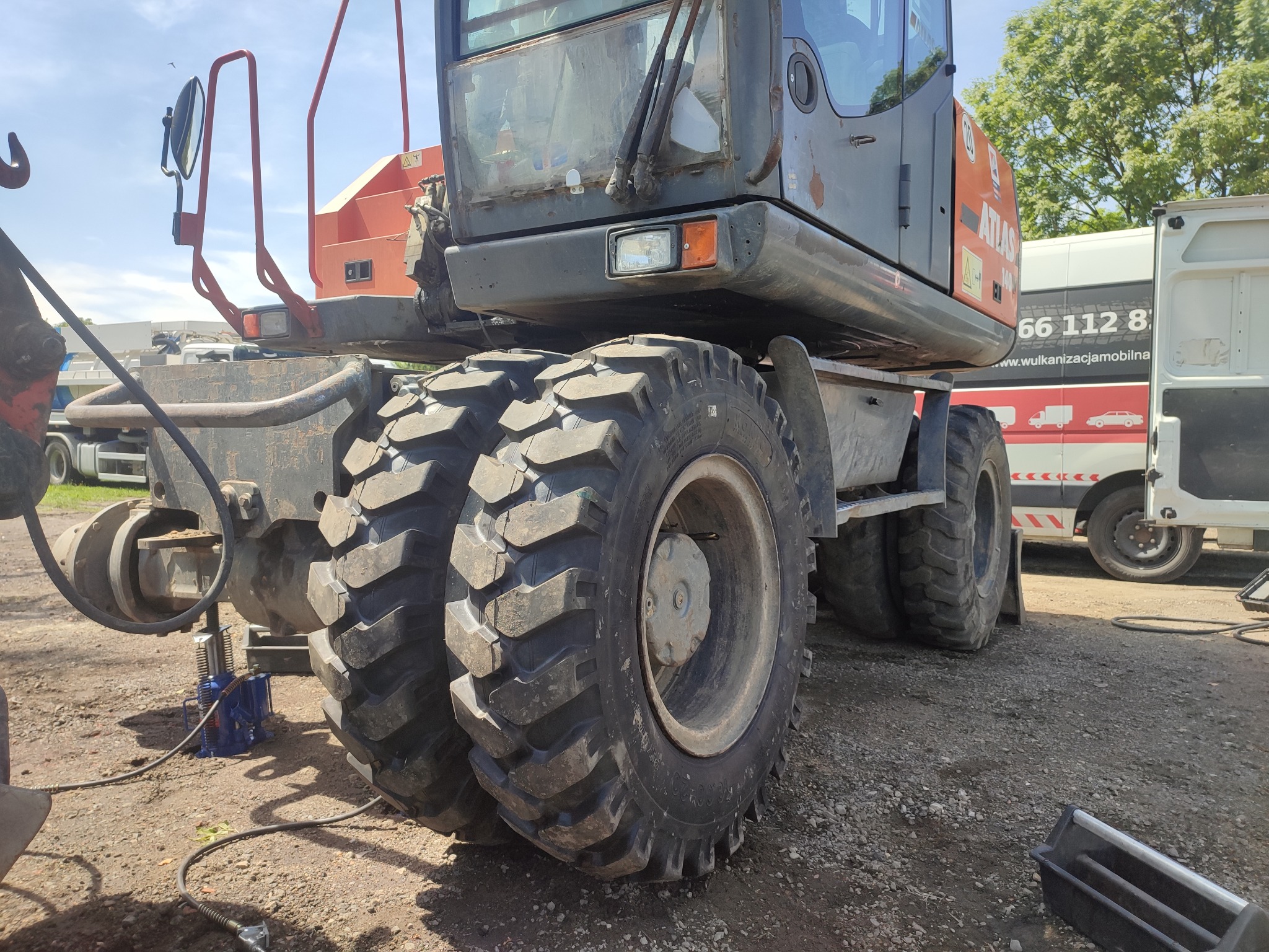 Koła koparki Atlas podnoszone za pomocą niebieskiego podnośnika hydraulicznego na żwirowym podłożu. W tle pojazd wulkanizacji mobilnej i drzewa.