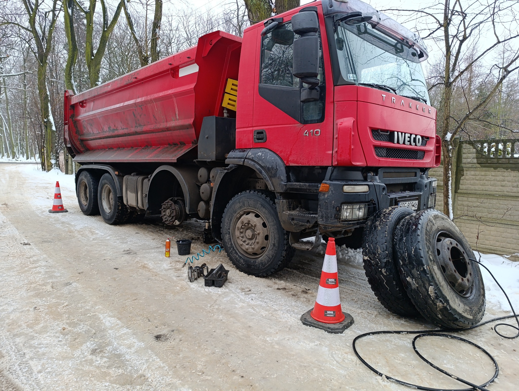 Czerwona ciężarówka Iveco Trakker z podniesioną kabiną podczas wymiany koła na drodze, widoczne narzędzia i koła obok, teren zimowy.