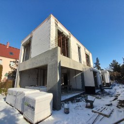 VERTE - Budowa domu z białych silikatów w zimowej scenerii. Widoczne ściany, betonowe elementy konstrukcyjne i palety z materiałem budowlanym. Słoneczny dzień, błękitne niebo.