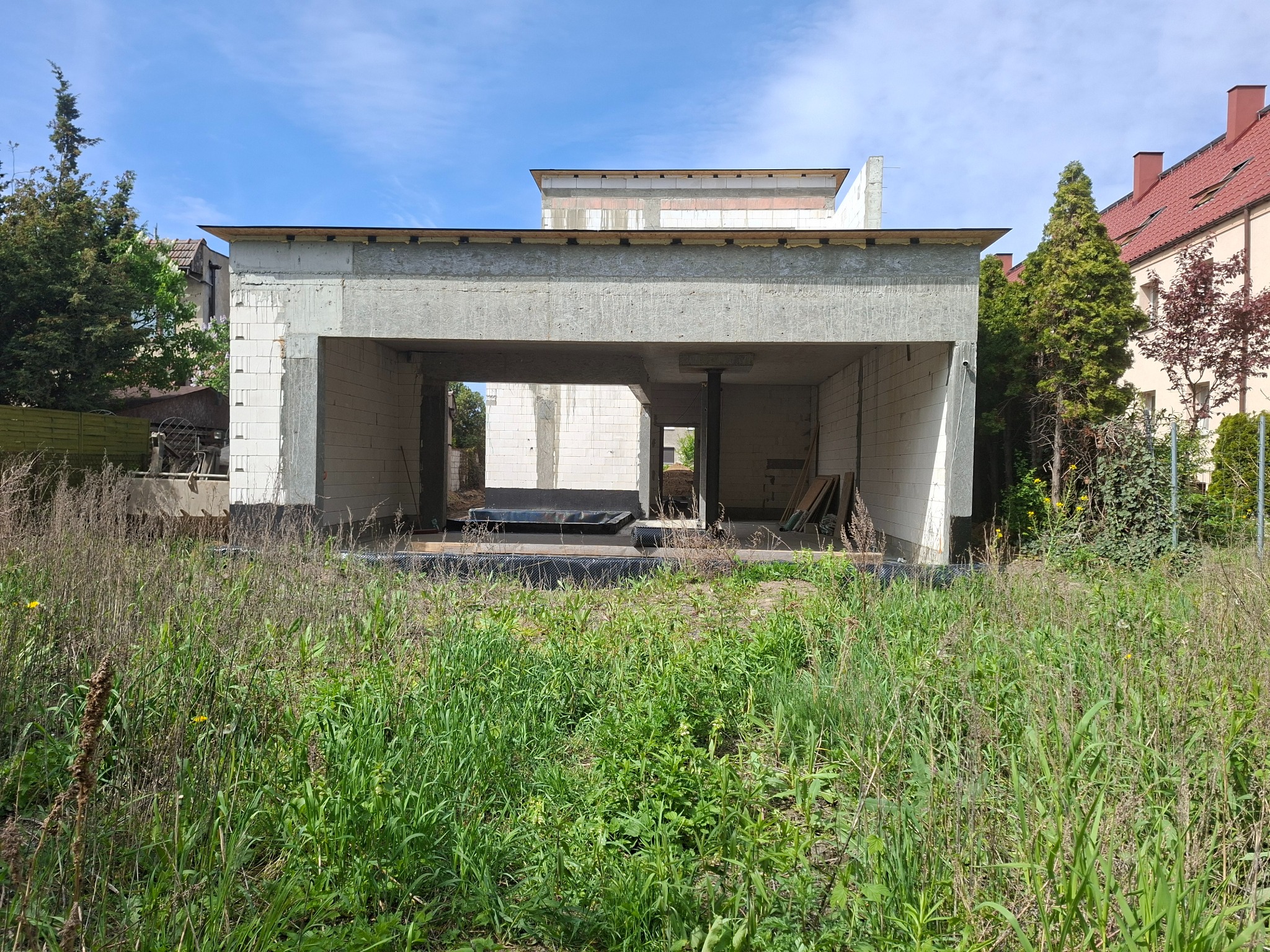 Surowy, otwarty szkielet domu z betonu komórkowego i żelbetu w otoczeniu zieleni. Widoczne elementy konstrukcyjne i nieukończone ściany. Budowa w toku.