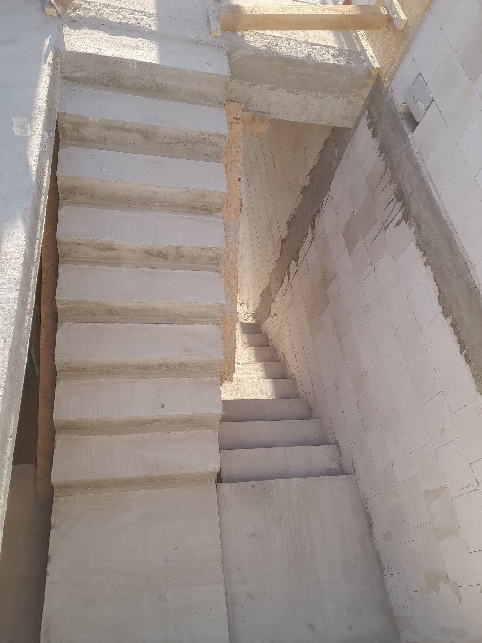 Wnętrze budynku w trakcie budowy. Widoczne surowe, betonowe schody prowadzące w dół, oraz fragment ściany z białych bloczków. Surowy stan wykończenia.