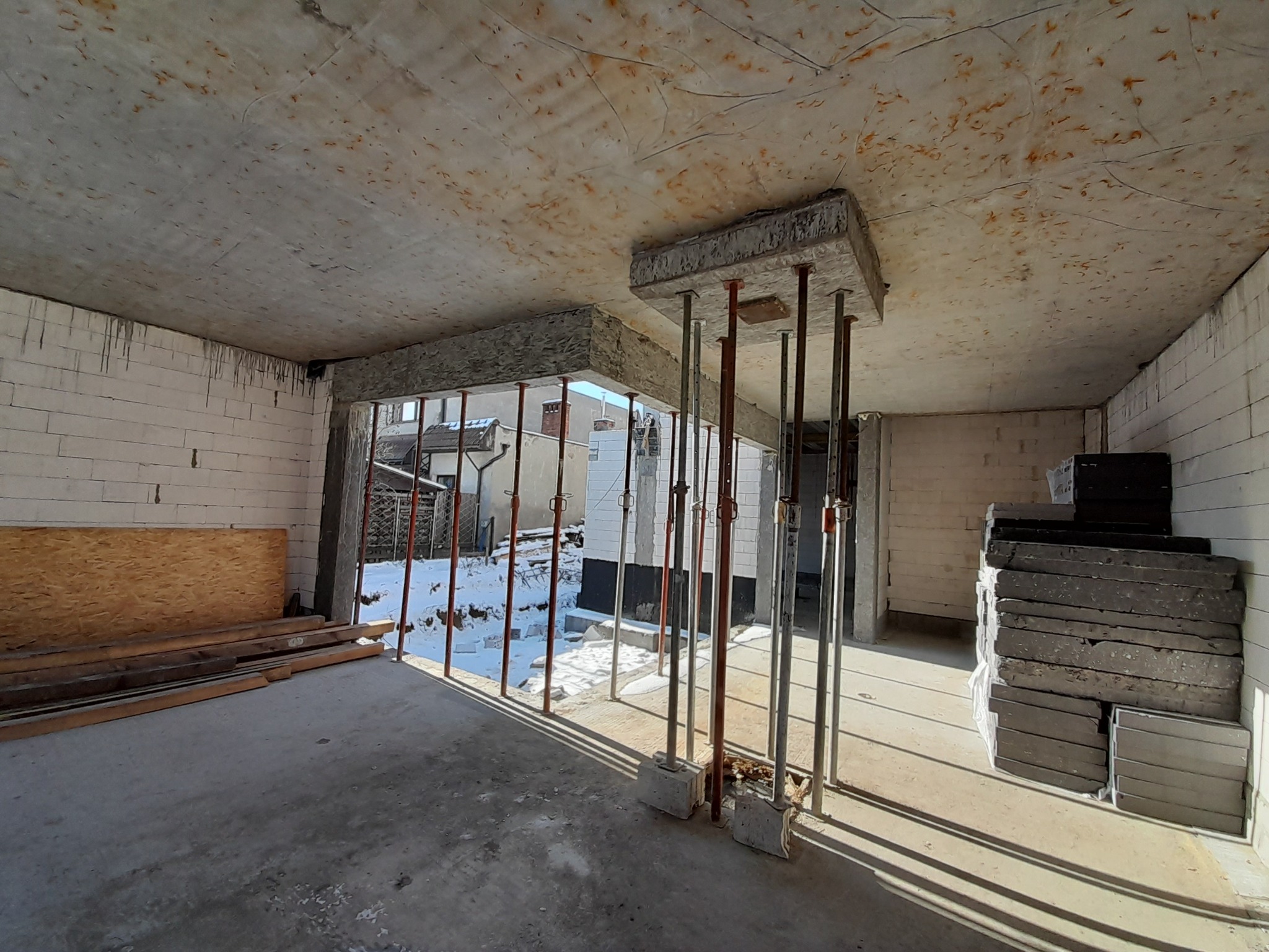 Wnętrze surowego budynku z podpieranym stropem betonowym. Widoczne pustaki, deski i podpory stropowe. Na zewnątrz śnieg. Wnętrze w stanie budowy.