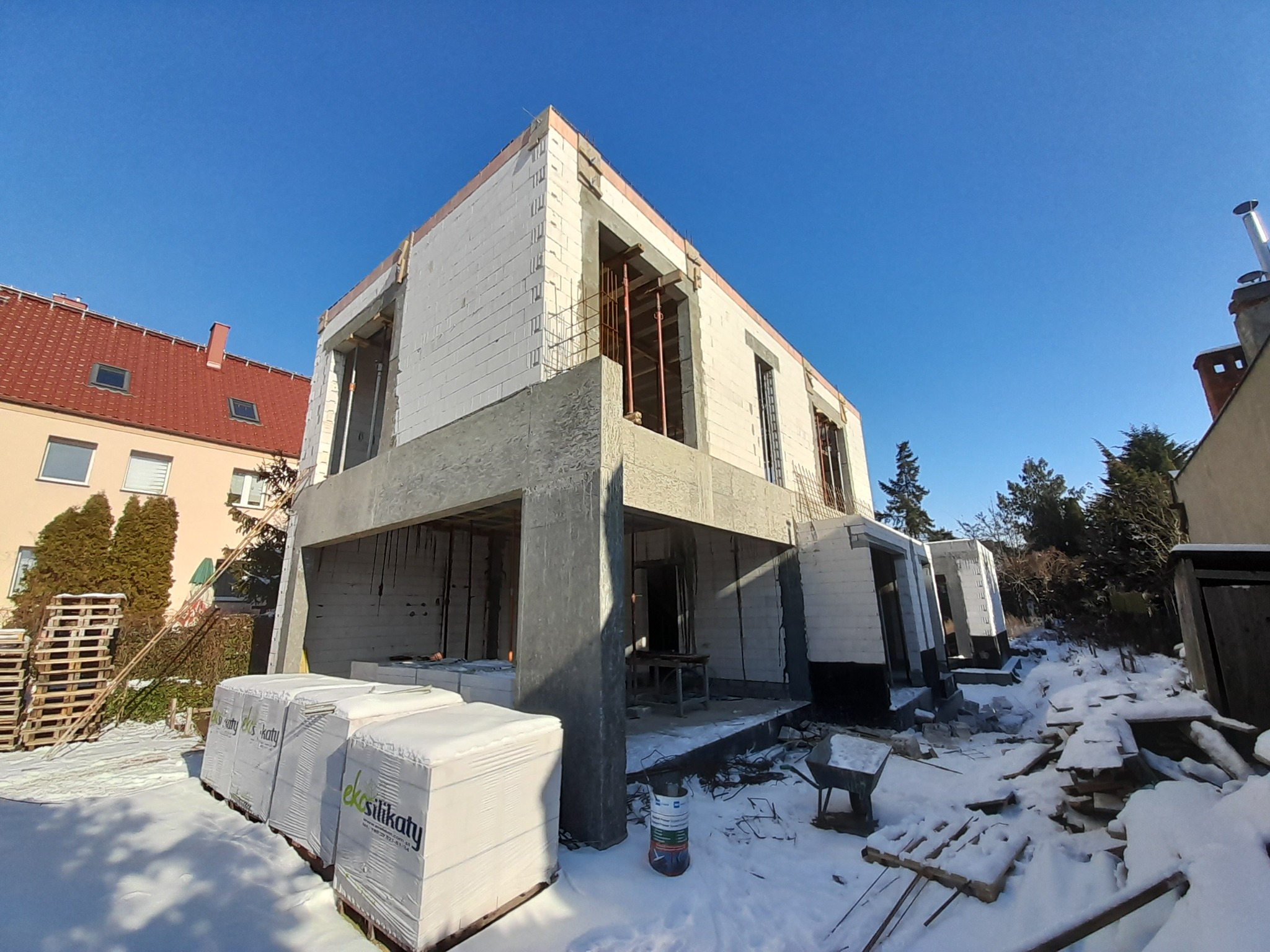 Budowa domu z białych silikatów w zimowej scenerii. Widoczne ściany, betonowe elementy konstrukcyjne i palety z materiałem budowlanym. Słoneczny dzień, błękitne niebo.