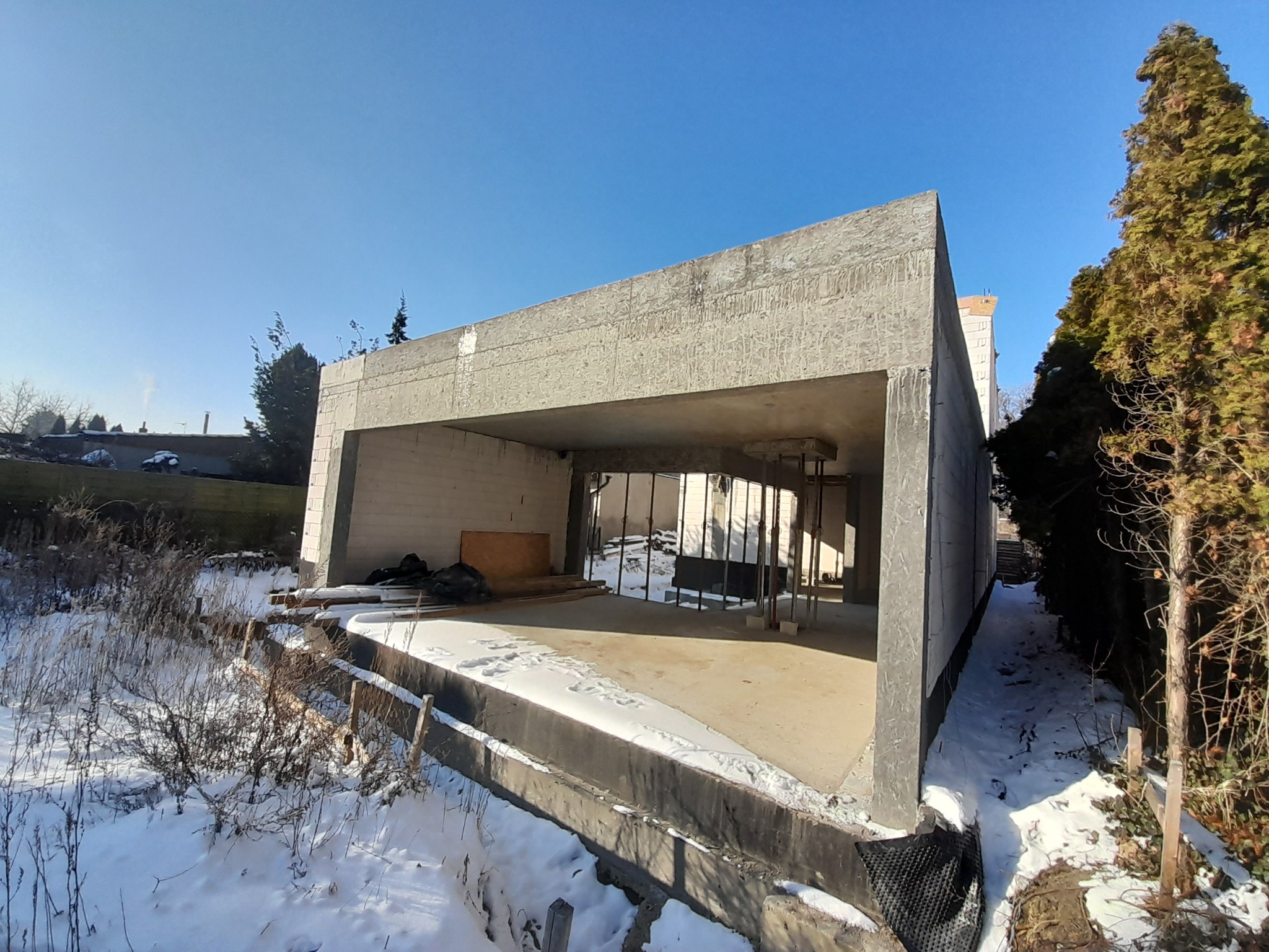 Surowy budynek żelbetowy w zimowej scenerii, z widoczną konstrukcją i elementami szalunku. Śnieg leży na fundamencie i w otoczeniu, podkreślając surowość i minimalizm.