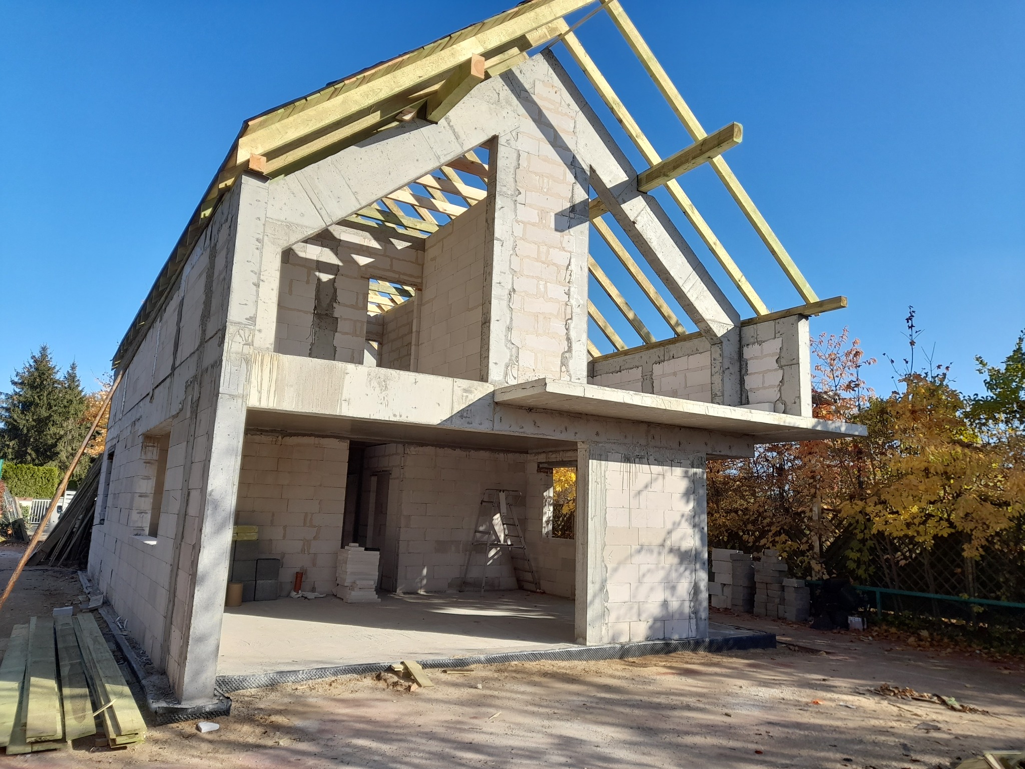 Surowy, dwukondygnacyjny dom z betonu komórkowego w trakcie budowy, z widoczną konstrukcją dachu z belek drewnianych na tle błękitnego nieba.