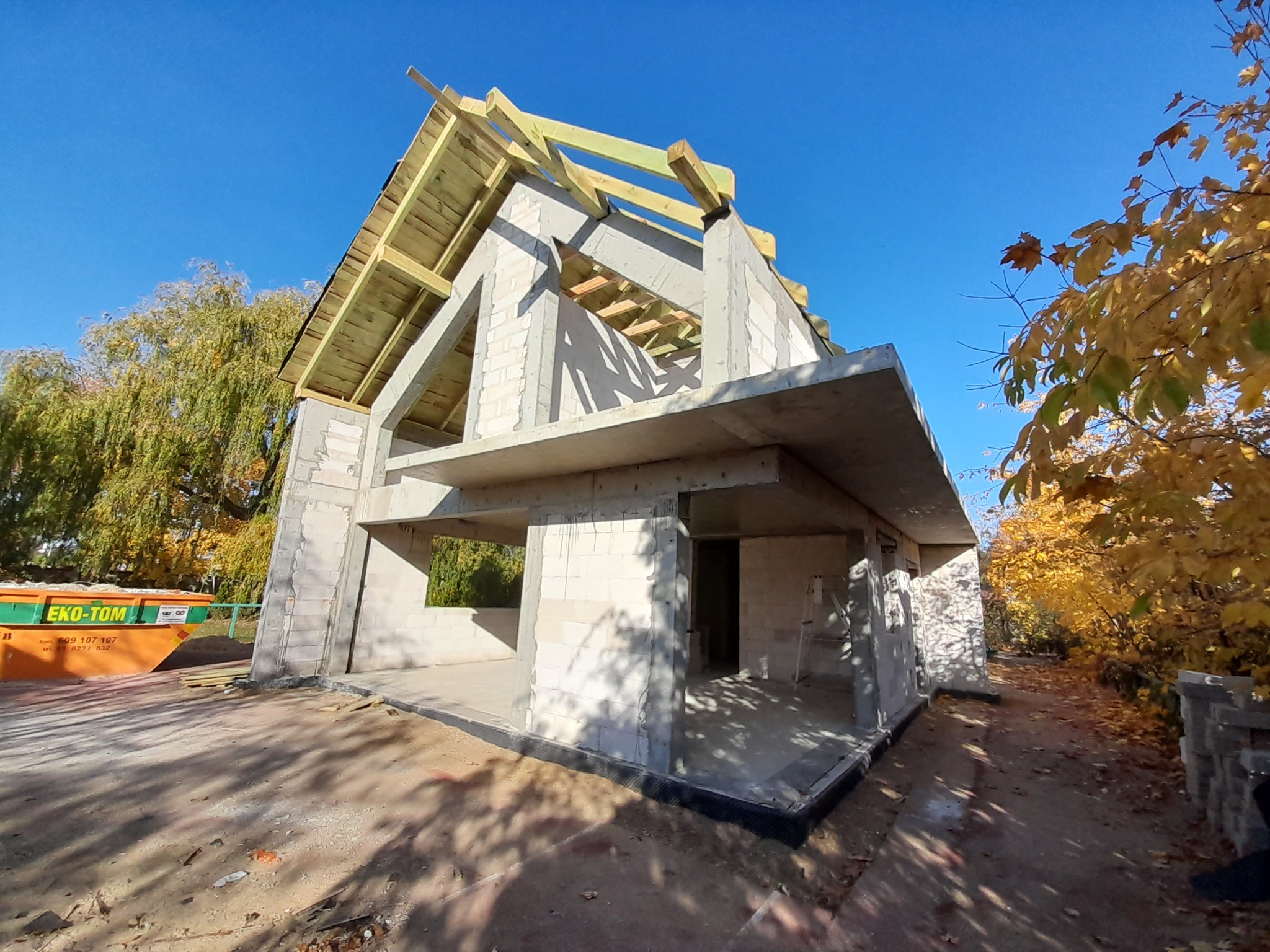 Surowy stan domu z bloczków, z widoczną konstrukcją dachu i elementami szalunku, na tle błękitnego nieba i drzew z jesiennymi liśćmi. Widoczny kontener budowlany.