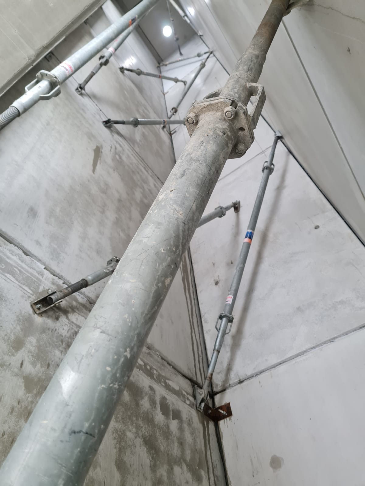 Szare, betonowe ściany podtrzymywane metalowymi stemplami budowlanymi. Widok z dołu, oświetlenie górne. Wnętrze w trakcie budowy.