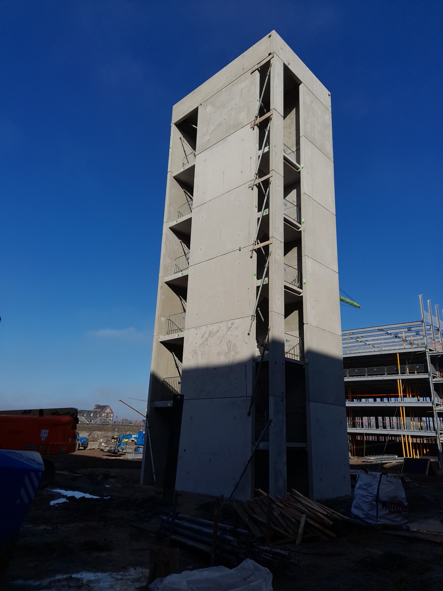 Surowy, wysoki budynek żelbetowy w trakcie budowy, z widocznymi elementami konstrukcyjnymi i otwartymi przestrzeniami balkonowymi na tle błękitnego nieba.