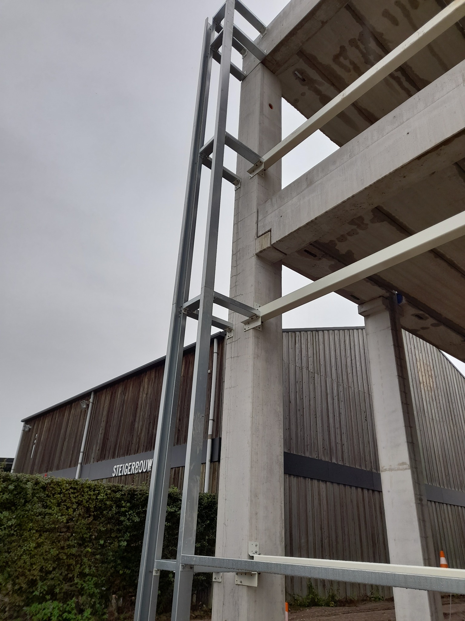 Surowa konstrukcja żelbetowa z szalunkiem na tle drewnianego budynku przemysłowego i pochmurnego nieba. Widoczne detale łączeń i surowy beton.