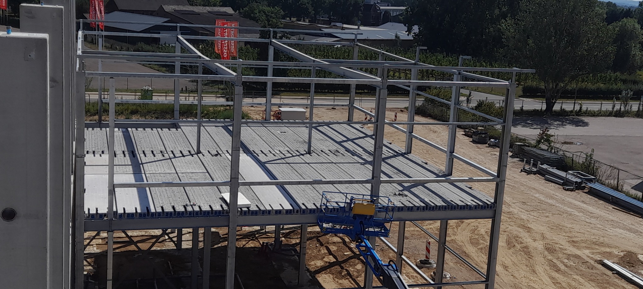 Wznoszenie konstrukcji hali stalowej z prefabrykowanych płyt stropowych. Widoczny podnośnik koszowy i elementy konstrukcyjne. Ujęcie z góry, w tle zabudowa i zieleń.