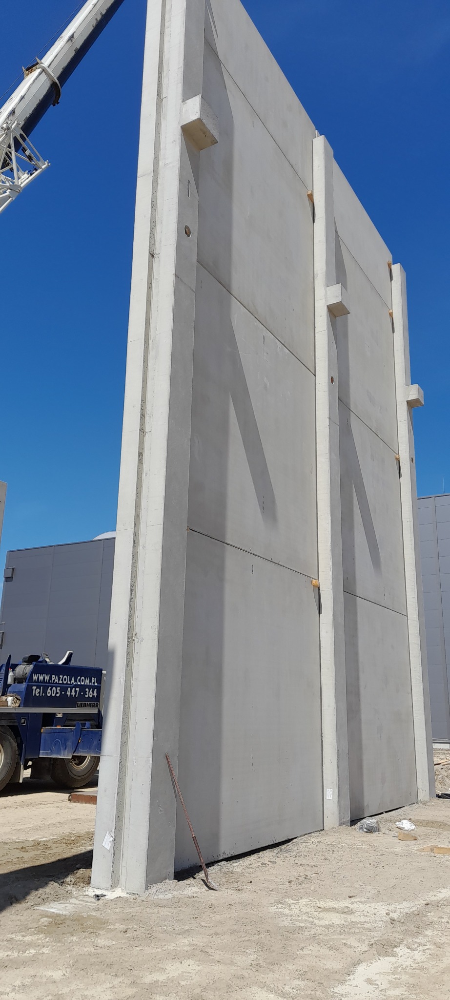 Montaż pionowej ściany z prefabrykatów betonowych w Poznaniu. Widoczny dźwig i transport, błękitne niebo, szara ściana z żelbetu, plac budowy.