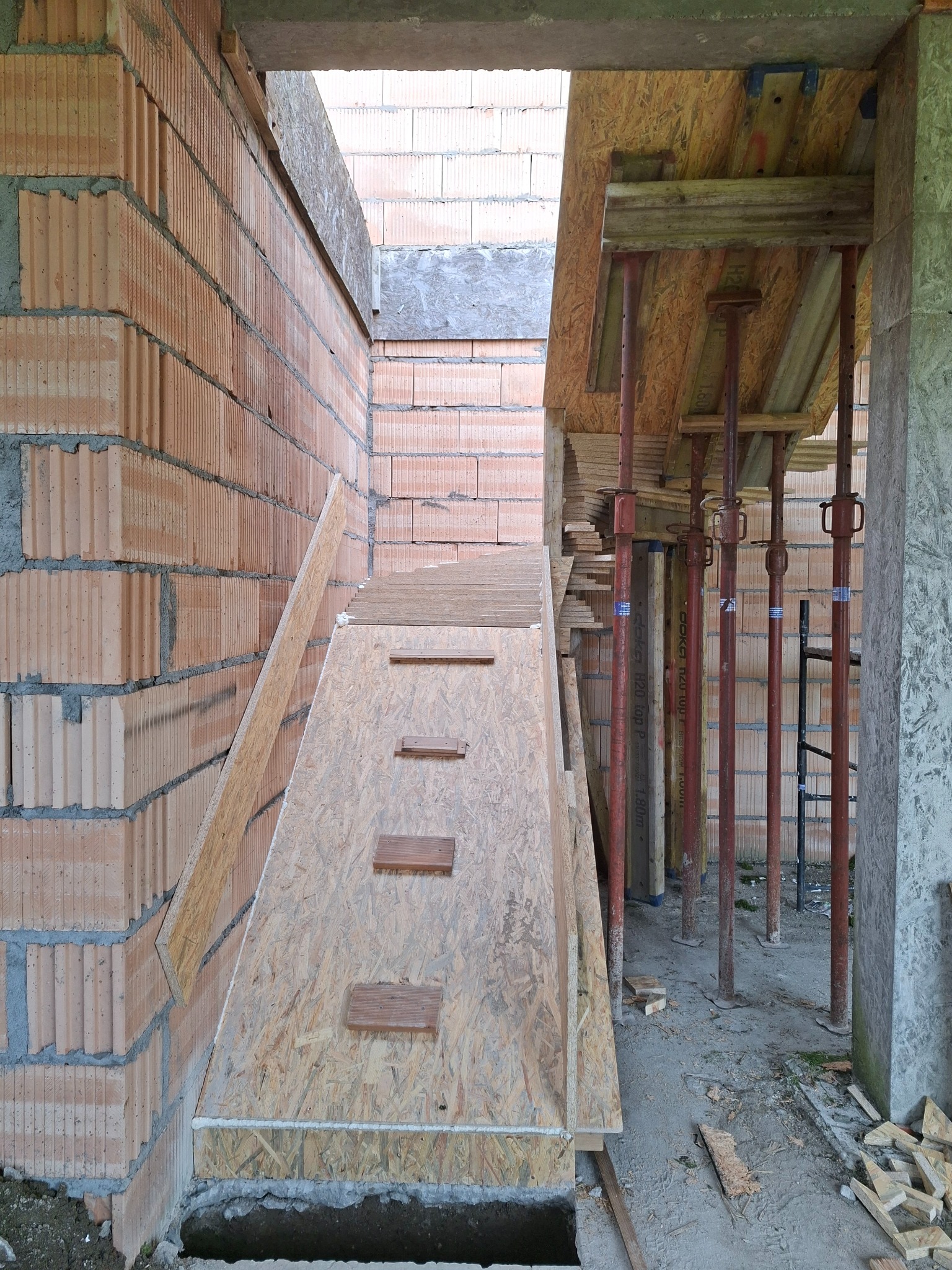 Wnętrze budowy: surowe mury z czerwonej cegły, drewniane szalunki pod schody, metalowe podpory stropu. Widoczny etap prac konstrukcyjnych.