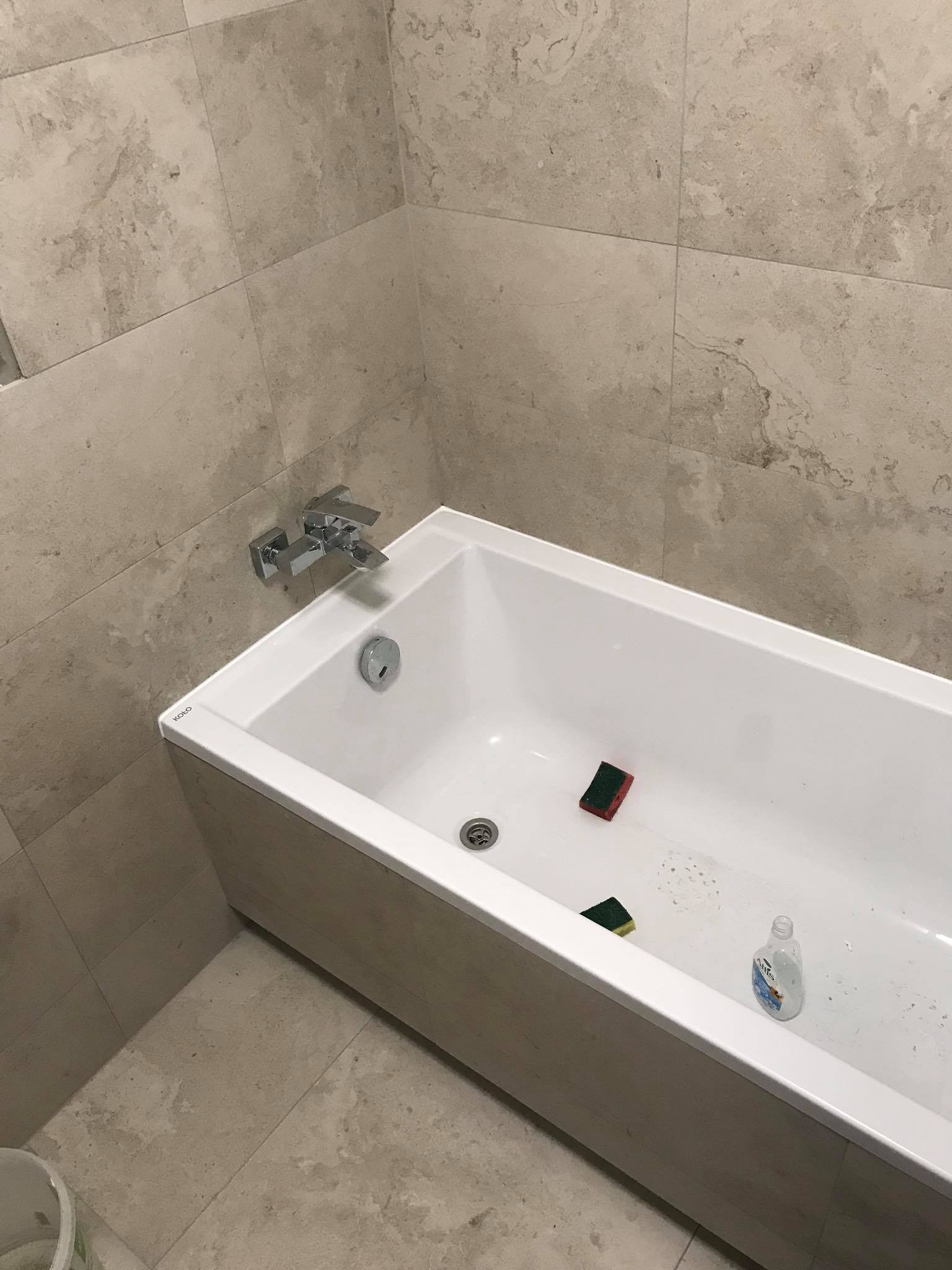 Biała wanna z chromowaną armaturą, wyłożona beżowymi płytkami imitującymi kamień. Widoczne gąbki i butelka płynu do czyszczenia. Ujęcie z góry, z bliska.