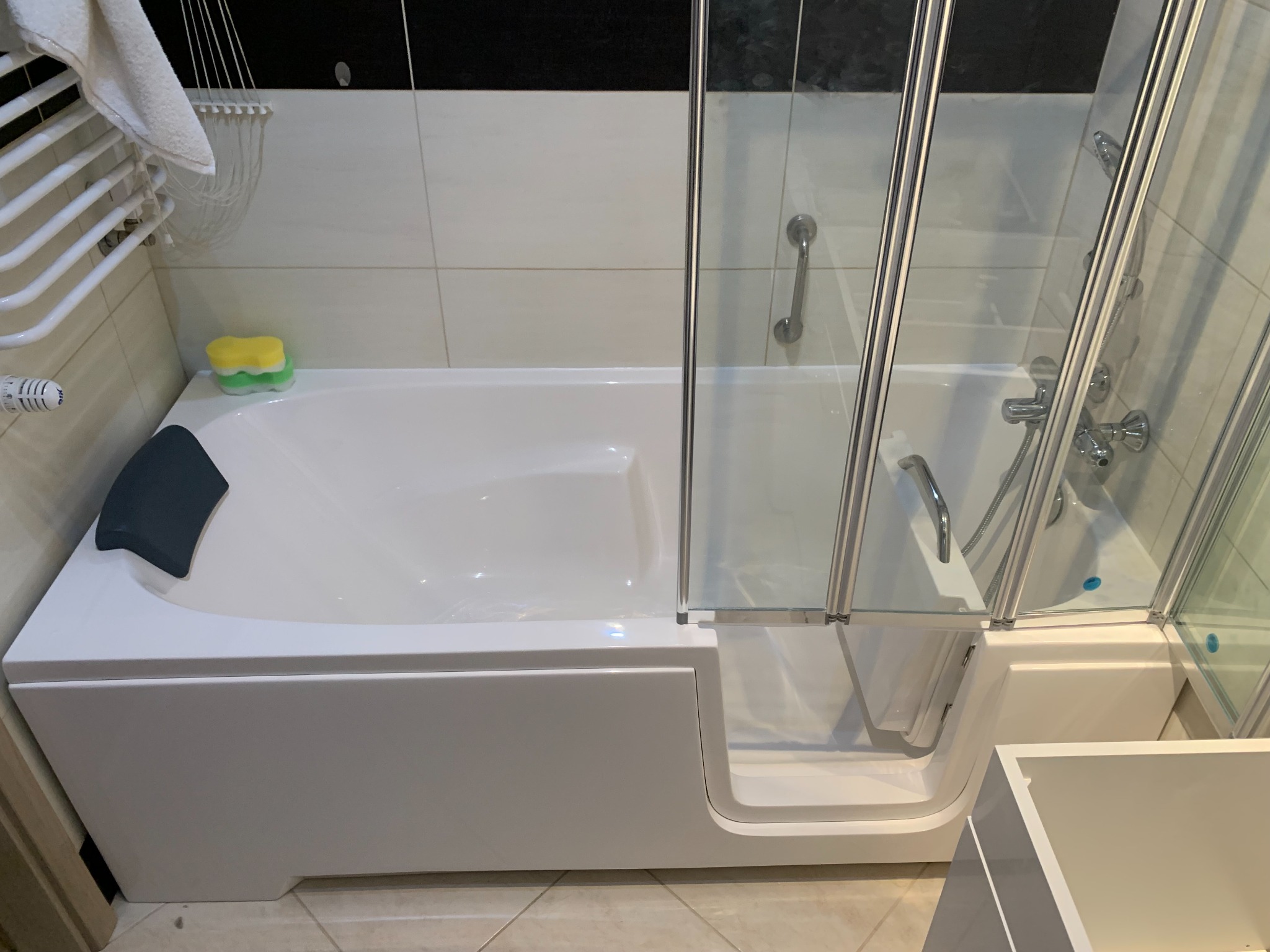 Biała wanna z hydromasażem i niskim wejściem, osłonięta szklanym parawanem. Widoczny grzejnik drabinkowy i fragment umywalki. Ujęcie w łazience z jasnymi płytkami.