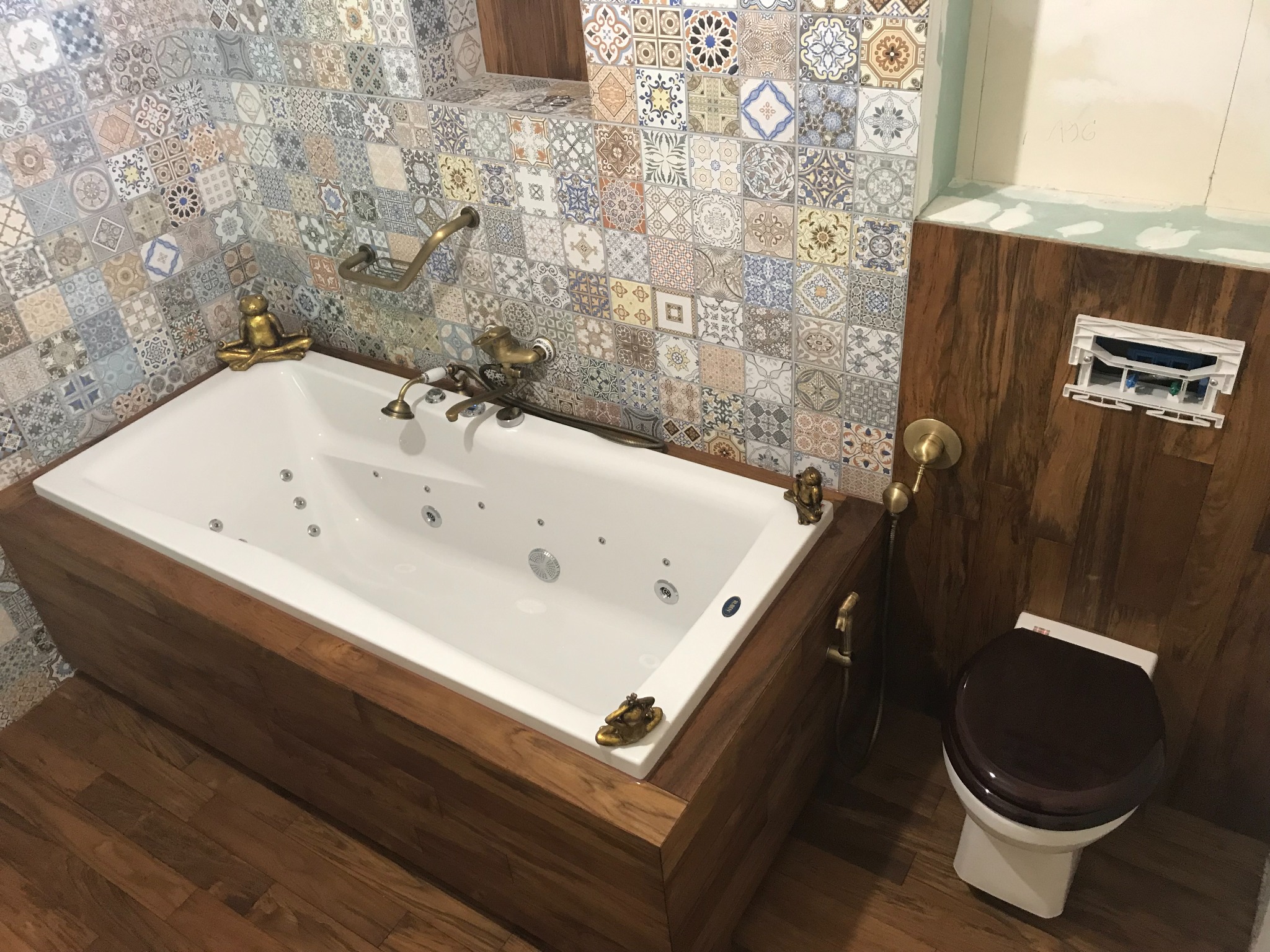 Wykończona łazienka z wanną jacuzzi obudowaną drewnem, mozaiką na ścianach i toaletą w stylu retro. Widoczne złote akcenty i figurki dekoracyjne.