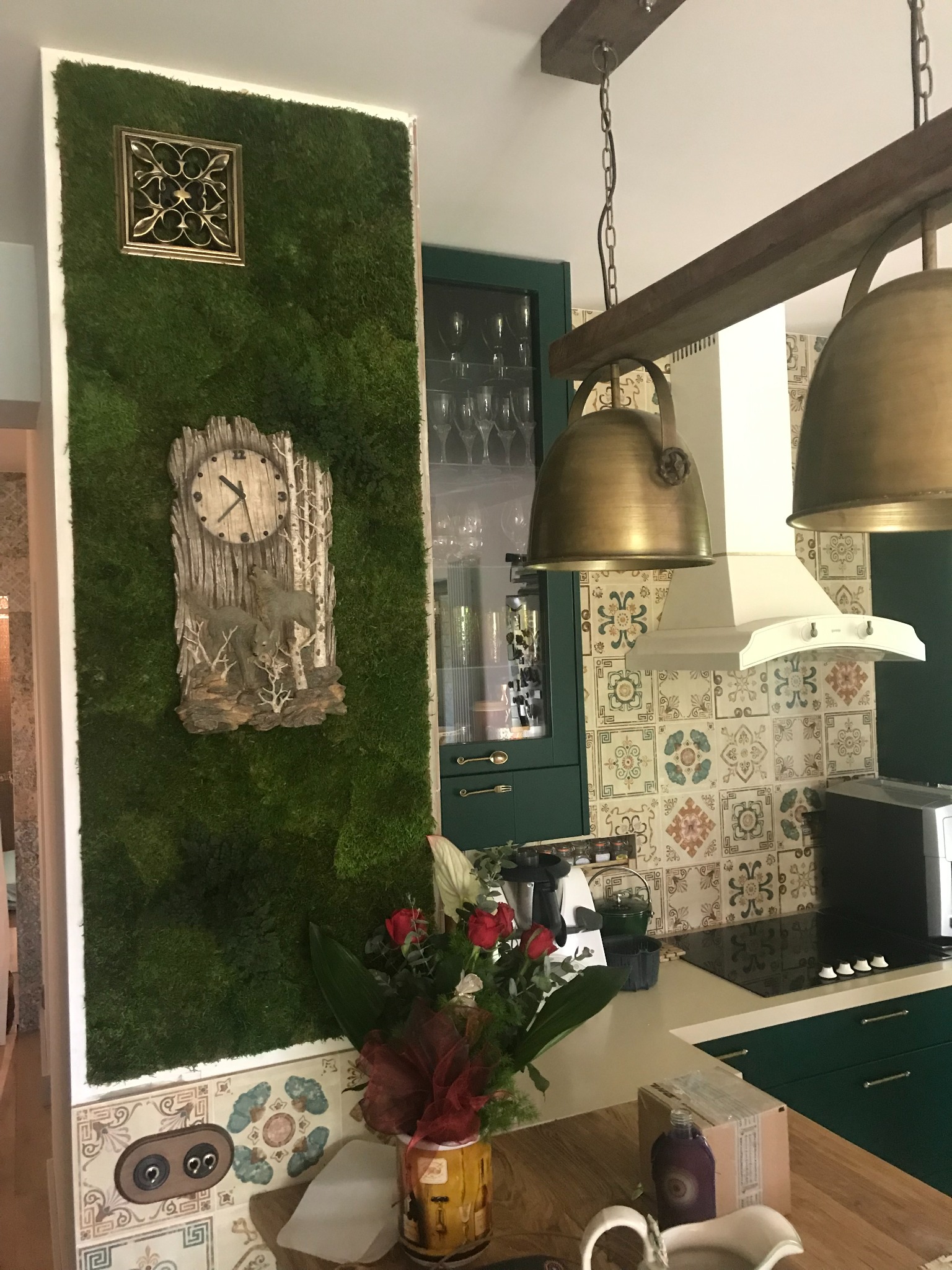 Kuchenna ściana z mchu dekoracyjnego, ozdobiona zegarem z motywem wilków i metalową kratką. W tle widoczne zielone szafki i mozaika na ścianie.