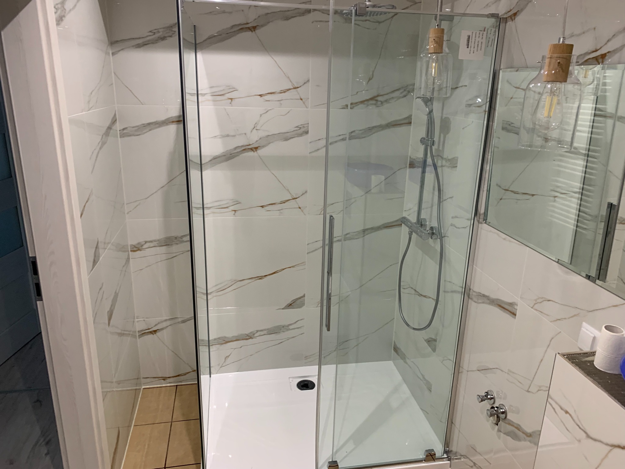 Nowoczesna kabina prysznicowa z białym brodzikiem i marmurowymi płytkami z brązowymi żyłami. Widoczne szklane drzwi, bateria i dekoracyjne żarówki.