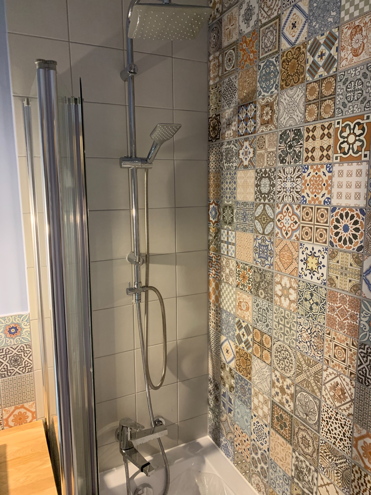 Wykończona łazienka z kabiną prysznicową i baterią, ściana wyłożona mozaiką z wzorzystych płytek w ciepłych odcieniach. Widoczny fragment blatu i biała wanna.