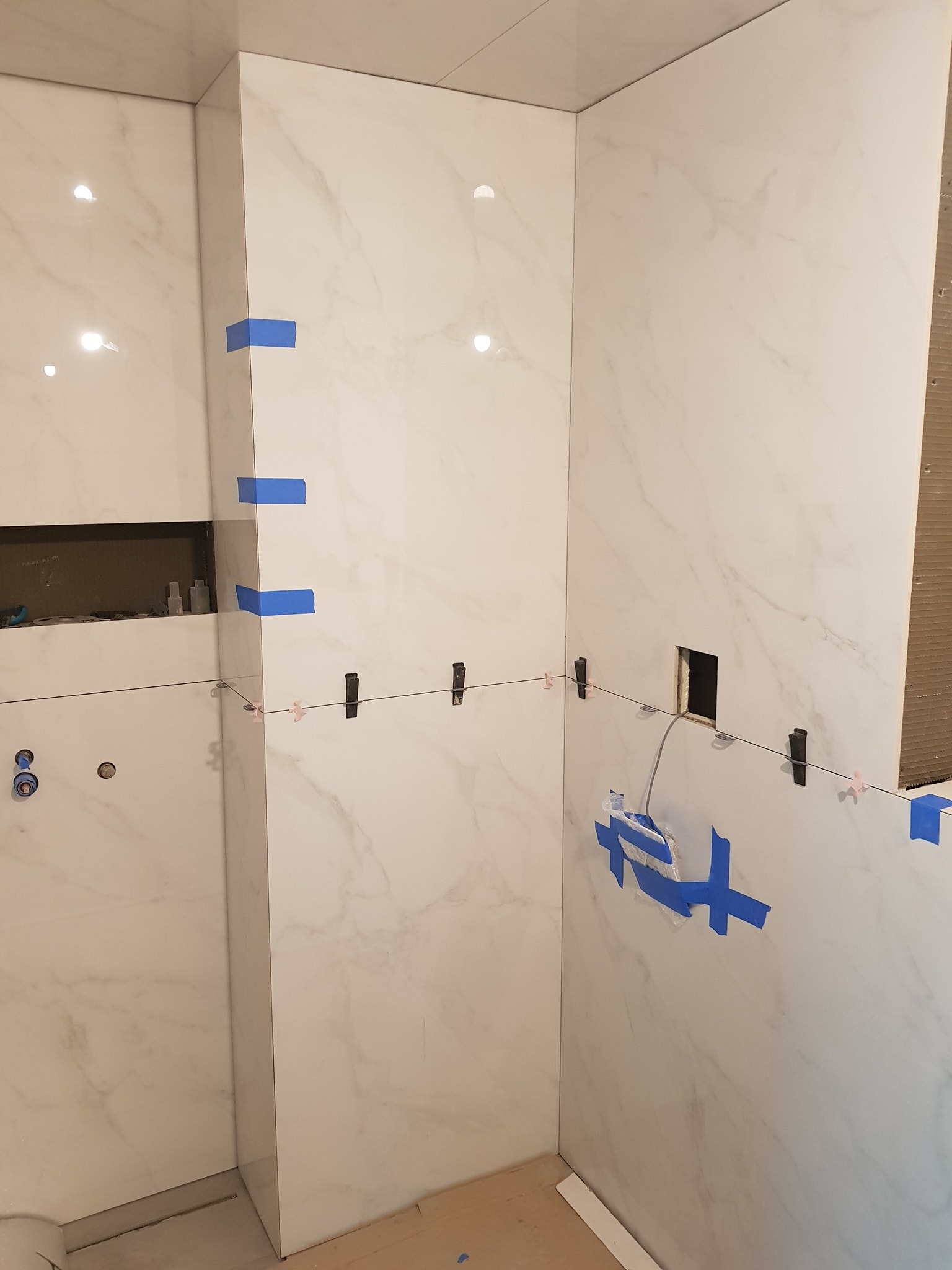 Ściany łazienki w trakcie układania białych płytek imitujących marmur, z użyciem niebieskiej taśmy i klipsów poziomujących. Widoczne otwory na instalacje.