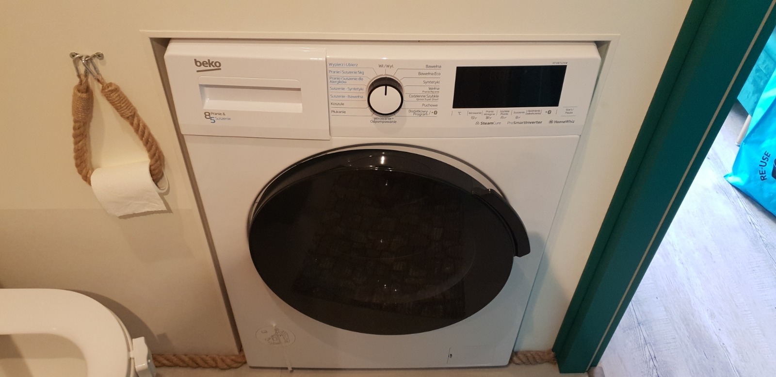 Biała pralka Beko w zabudowie łazienki w Łodzi. Obok toaleta i ozdobny uchwyt na papier toaletowy z liny jutowej. Fragment podłogi z szarymi panelami.