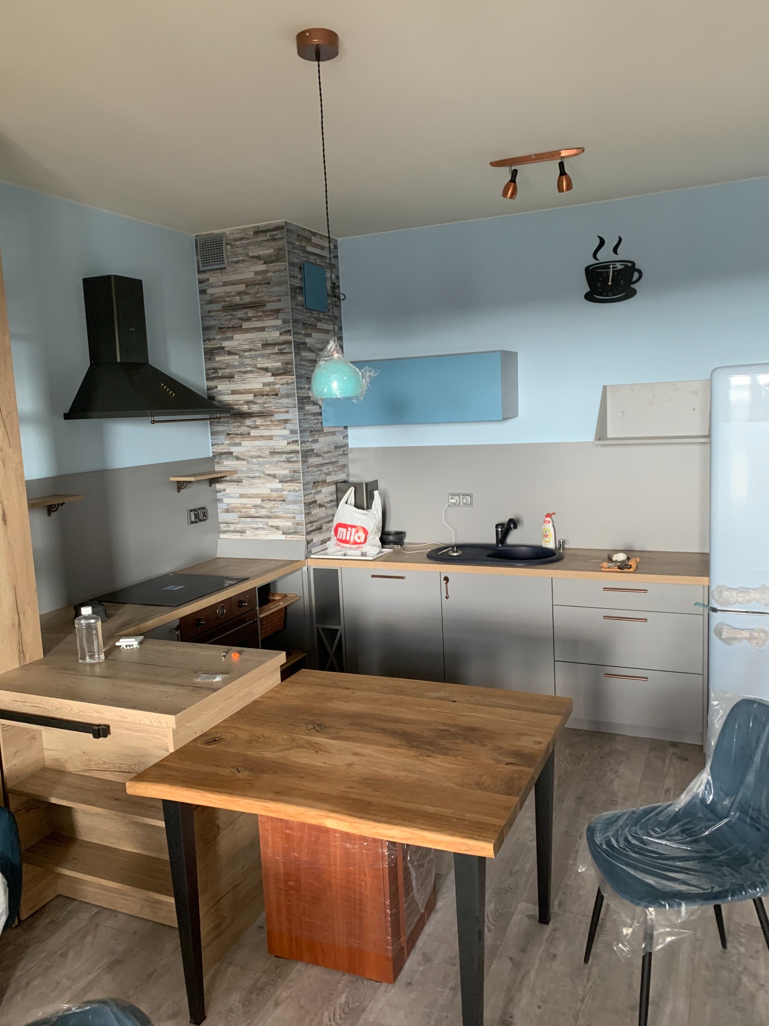 Jasna kuchnia z szarymi szafkami, drewnianym blatem i stołem. Na ścianie dekoracja filiżanki kawy. Krzesło w folii ochronnej, sugerujące świeży montaż.