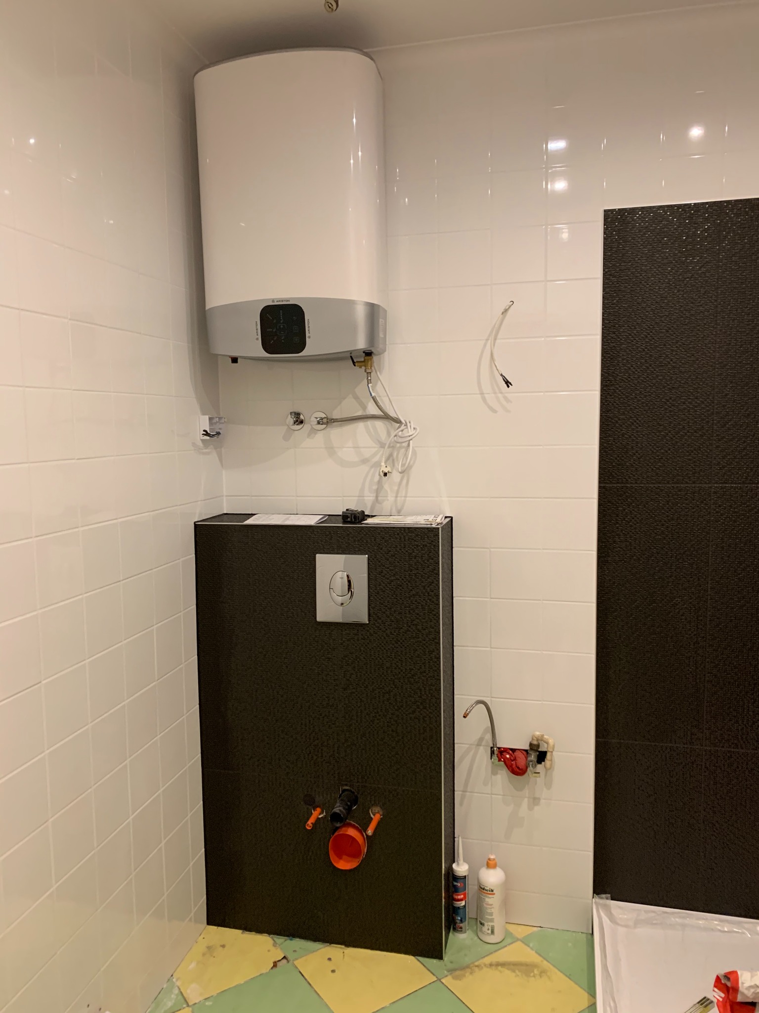 Remont łazienki w trakcie: Geberit z czarną mozaiką, bojler Ariston i białe płytki. Widoczne przyłącza hydrauliczne i akcesoria montażowe, podłoga częściowo wyłożona płytkami.