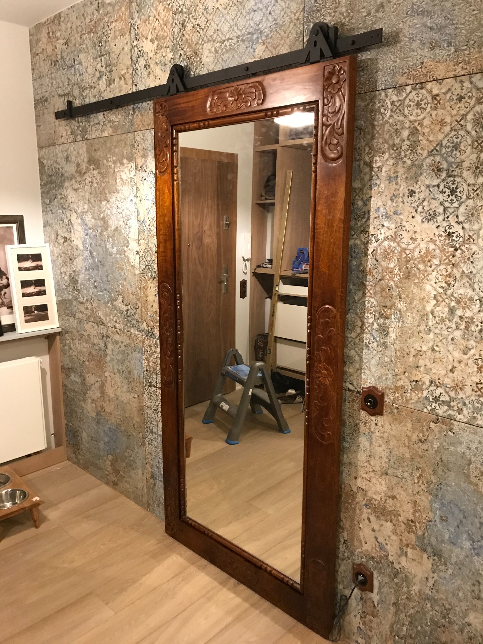 Rustykalne lustro przesuwne z dekoracyjną ramą, zawieszone na tle ściany wykończonej wzorzystymi płytkami w odcieniach beżu i szarości. Widoczna drabina i fragment podłogi.