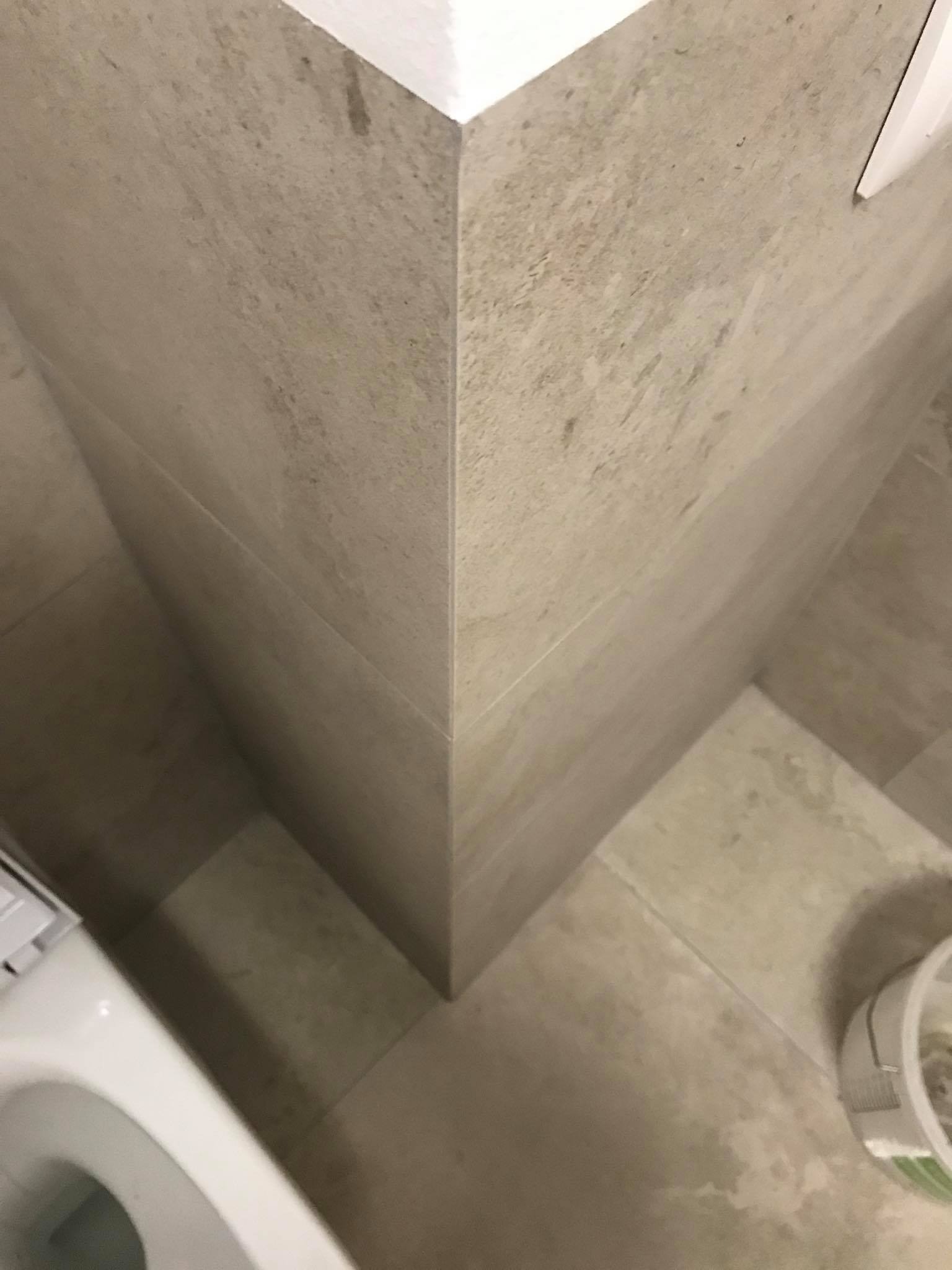 Szczegółowe ujęcie narożnika wykończonej łazienki z beżowymi płytkami, widoczny fragment toalety i wiaderko, podkreślające precyzję wykonania. Minimalistyczny design.