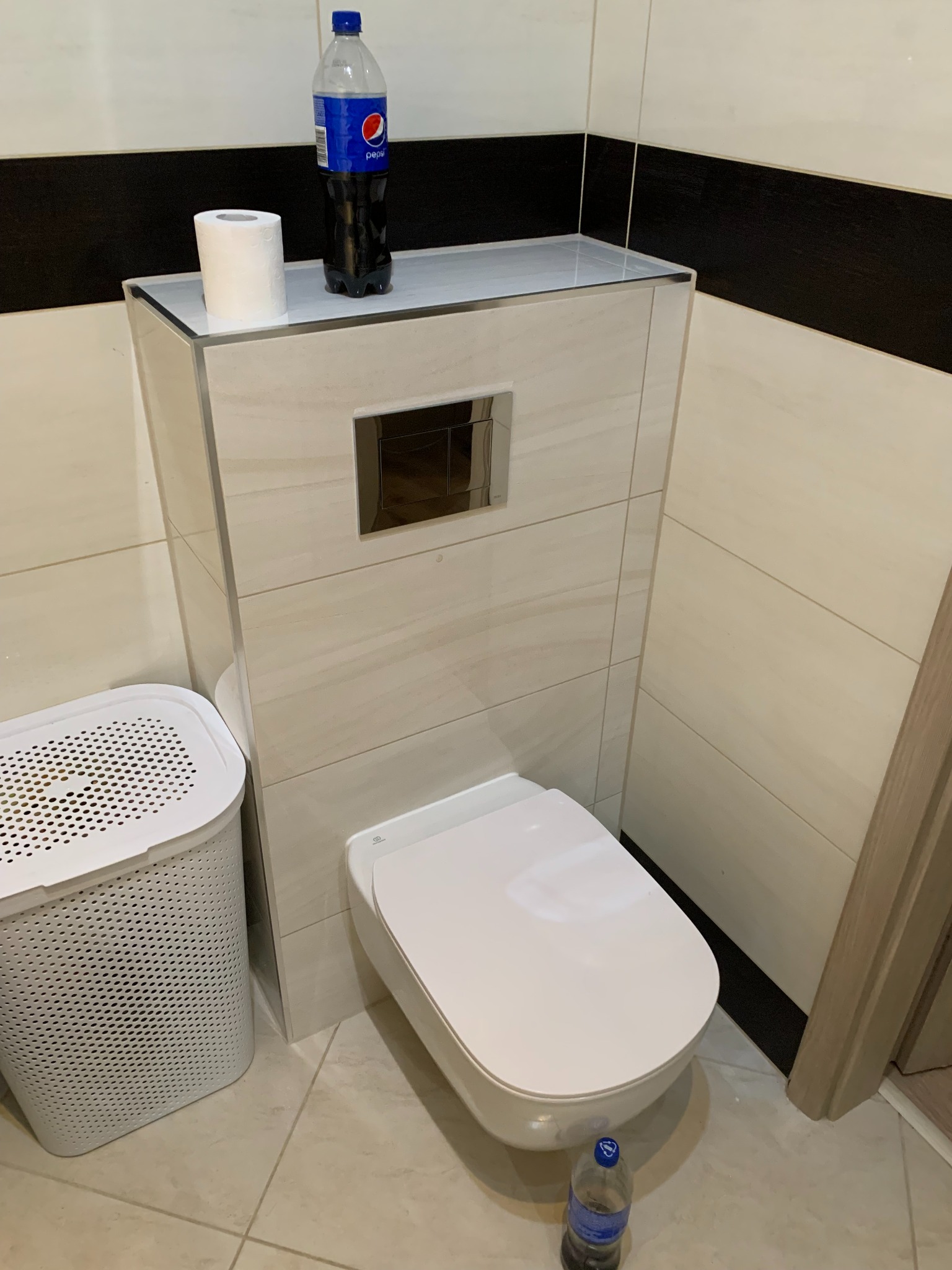 Nowoczesna toaleta podwieszana z zabudową z płytek imitujących marmur. Na zabudowie papier toaletowy i butelka Pepsi. Kosz na pranie obok. Ujęcie z dołu.