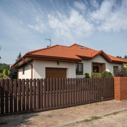 MPartners Nieruchomości Mariusz Michalski - Jednorodzinny dom z czerwoną dachówką, białymi ścianami i brązowym ogrodzeniem. Widok z zewnątrz w słoneczny dzień.