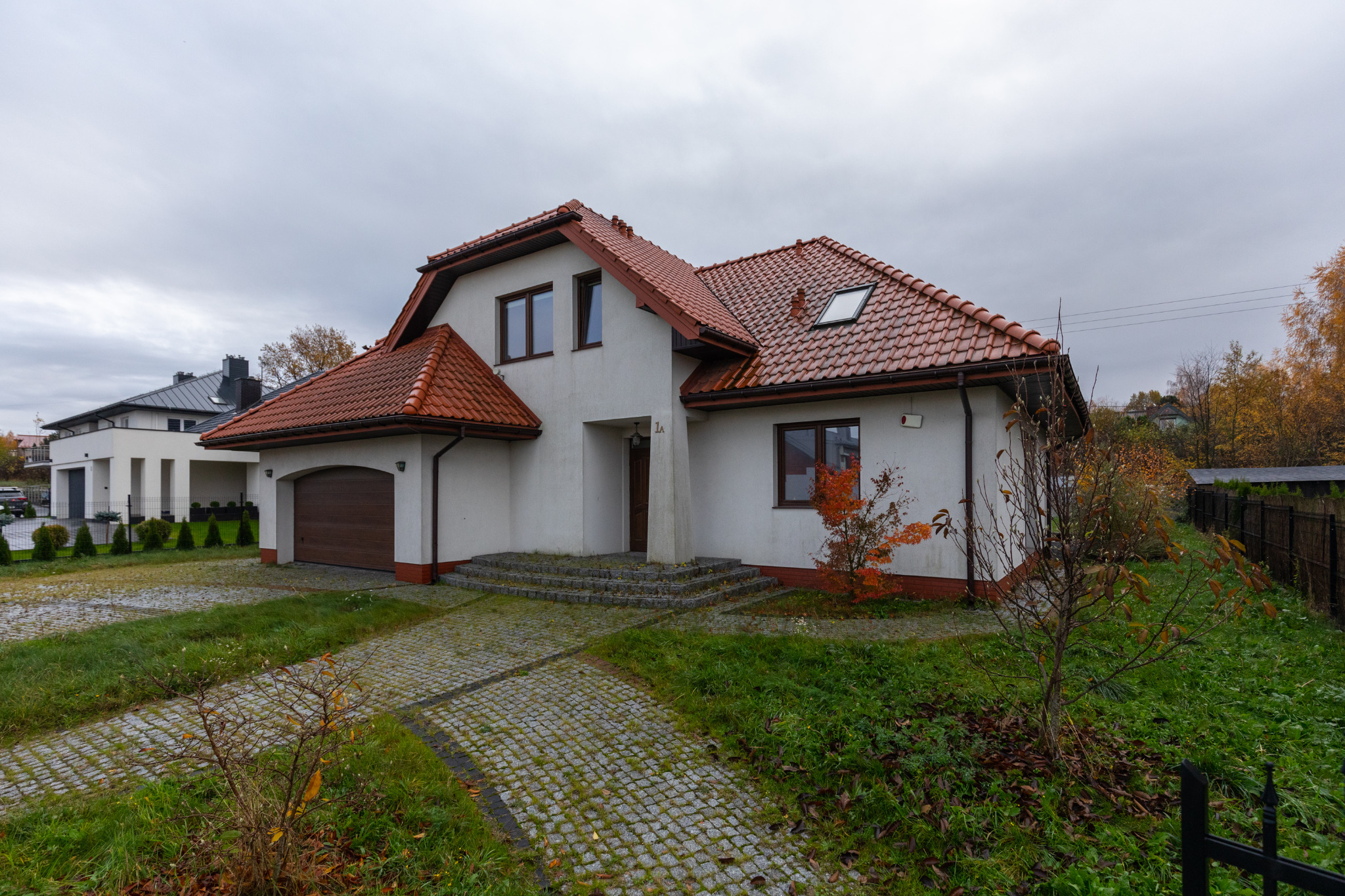 Biały dom z czerwoną dachówką i brązowymi oknami. Podjazd z kostki brukowej, trawnik. Widok z przodu na dom z numerem 1A. Pochmurne niebo w tle.