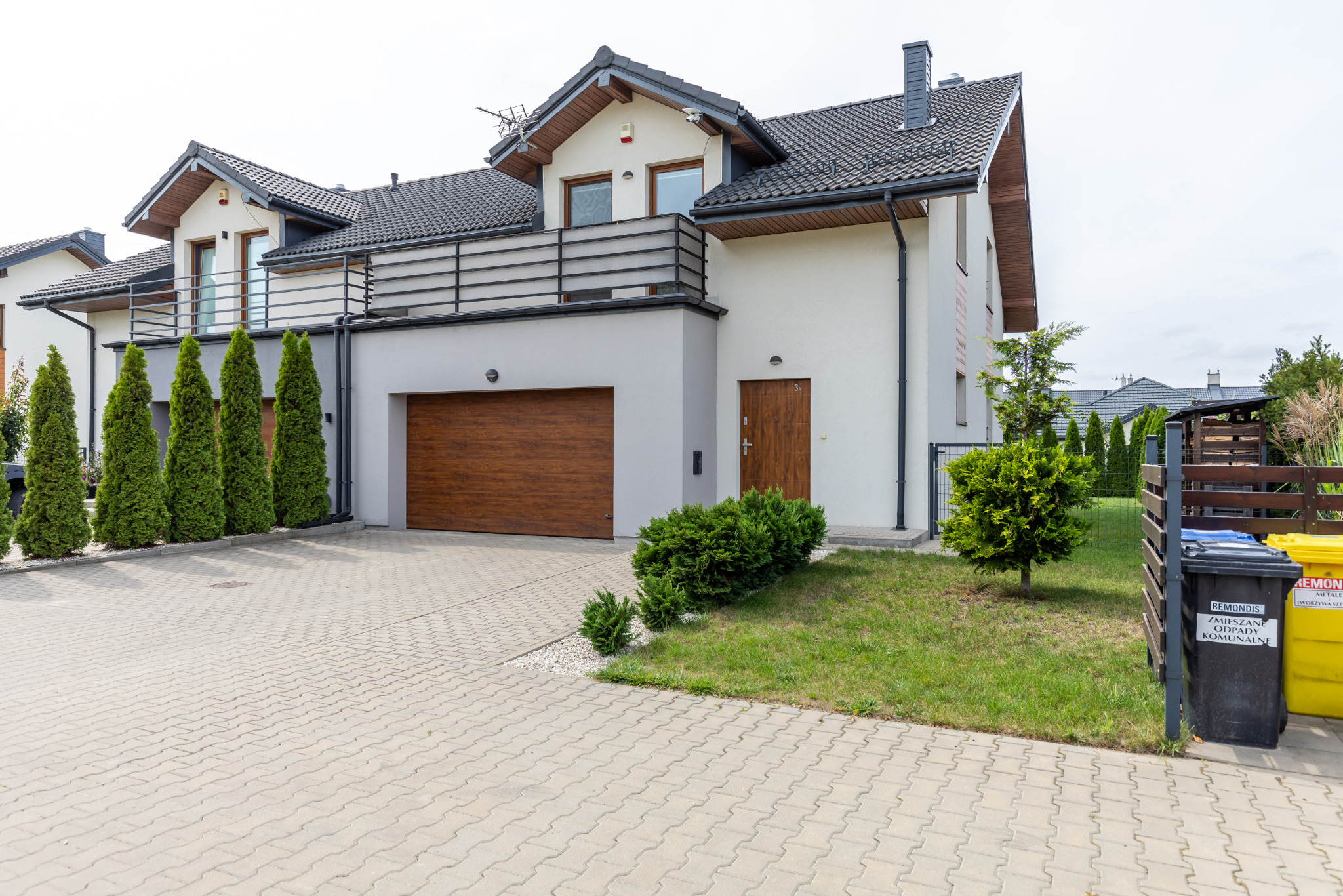 Nowoczesny dom szeregowy z balkonem i garażem, jasna elewacja, brązowe drzwi i brama, zadbany trawnik, kostka brukowa, pojemniki na śmieci. Prezentacja nieruchomości.