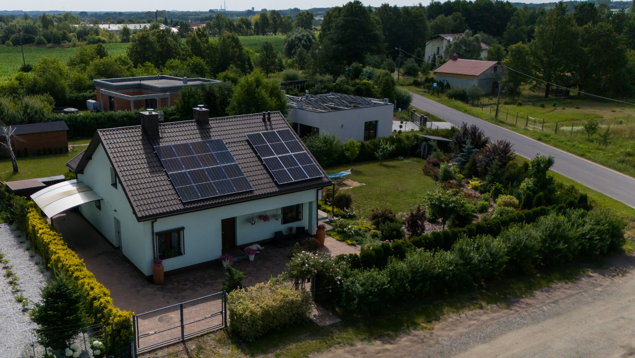 Dom z panelami słonecznymi na dachu, otoczony zielonym ogrodem i innymi budynkami w oddali. Ujęcie z lotu ptaka prezentuje nieruchomość.